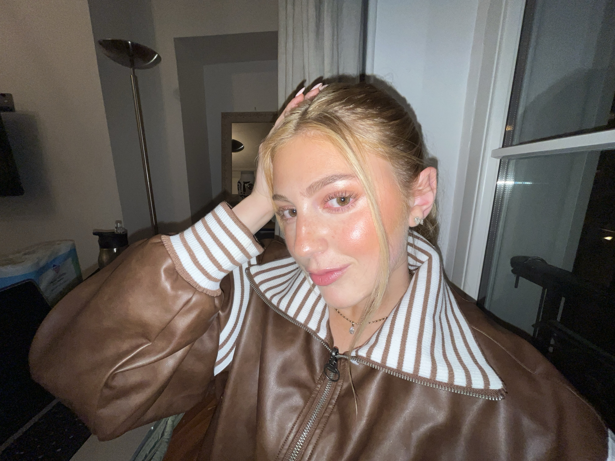 The image shows a young woman with blonde hair and brown eyes, wearing a brown leather jacket with white and brown striped collar and cuffs. She is touching her hair with her right hand. She is wearing a delicate necklace. Behind her, there is a window through which a nighttime urban scene is visible. The photo appears to have been taken indoors, with a lamp visible in the background. There are no specific visual indicators that place the image in Paris, France.