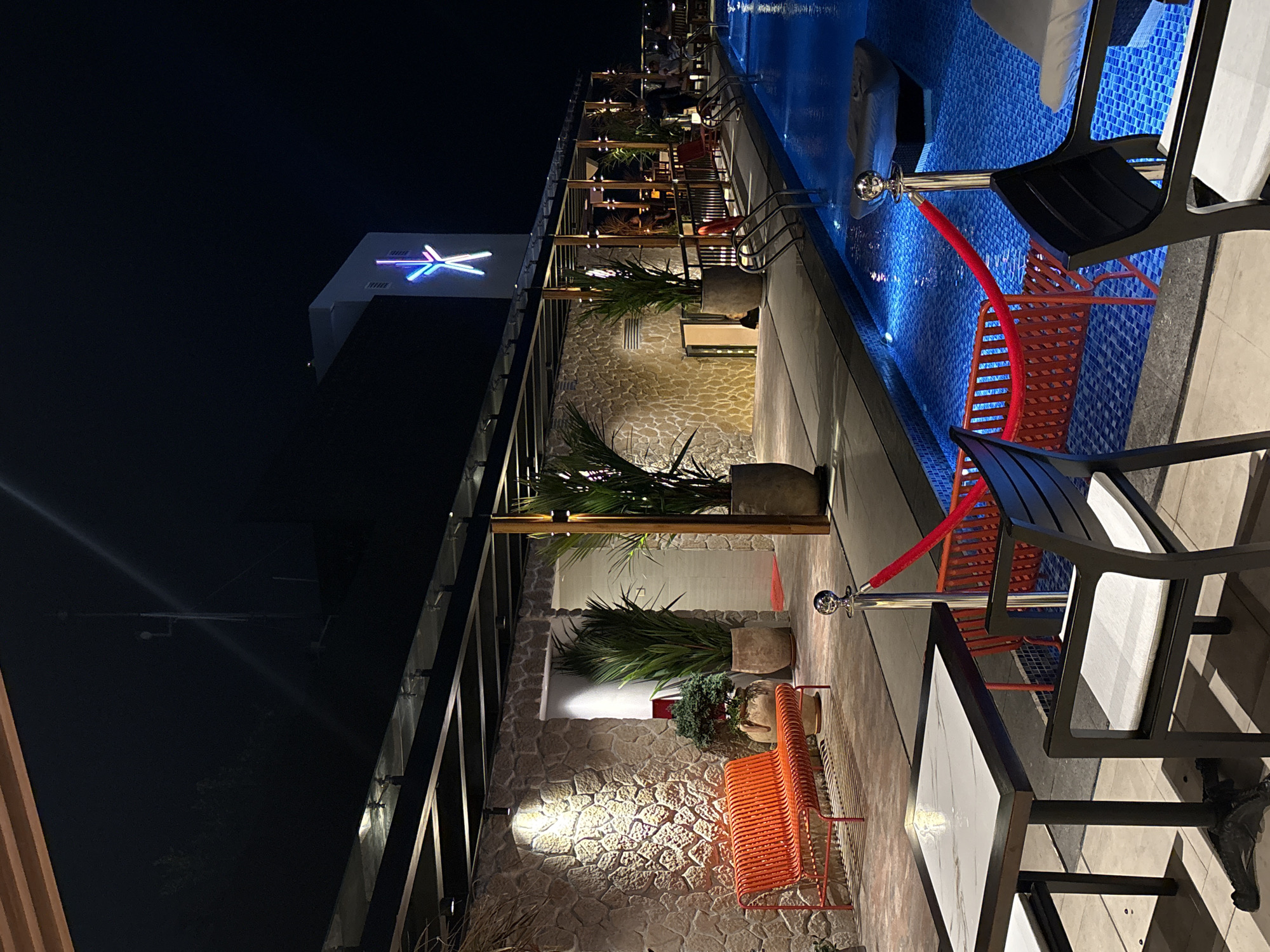 Captured at night in Nha Trang ward, Vietnam, the image features an outdoor dining area next to a pool illuminated with blue lights. A building with a neon logo is visible against the dark sky. Tables and chairs are arranged along the side of the pool, with plants in large pots separating sections. Red velvet ropes cordon off certain areas. The architectural style incorporates stone walls and a modern, open-air design.