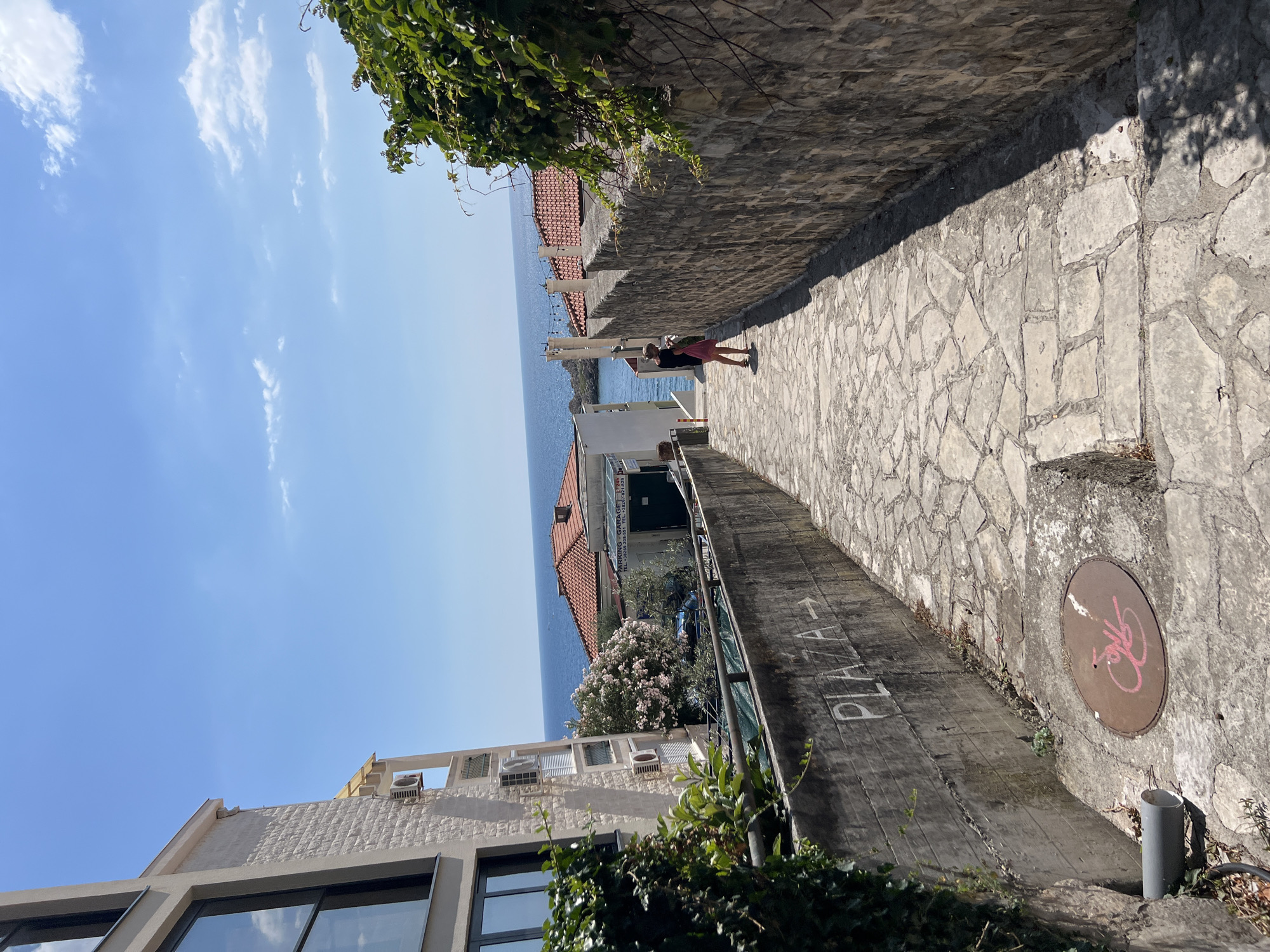 The image depicts a narrow, stone-paved walkway with a high stone wall on the right and buildings and the sea on the left. The walkway is bathed in sunlight, suggesting it is daytime, and the sky is a clear blue with a few wispy clouds. Green ivy grows along the top of the wall and the edges of the buildings.
In the middle distance, a person wearing a pink skirt and a dark top walks away from the camera down the path. Beyond them, the deep blue sea stretches to the horizon, meeting the clear sky. Red-tiled roofs of buildings are visible along the coast.
On the concrete retaining wall to the left of the walkway, the word "PLAZA" is stenciled in white with an arrow pointing forward. Further down the wall, a rusty circular manhole cover with a pink symbol is visible. A modern building with large windows and air conditioning units is present on the far left of the frame, partially obscured by greenery. The overall scene evokes a sense of a warm, sunny day in a coastal town, possibly during the summer.