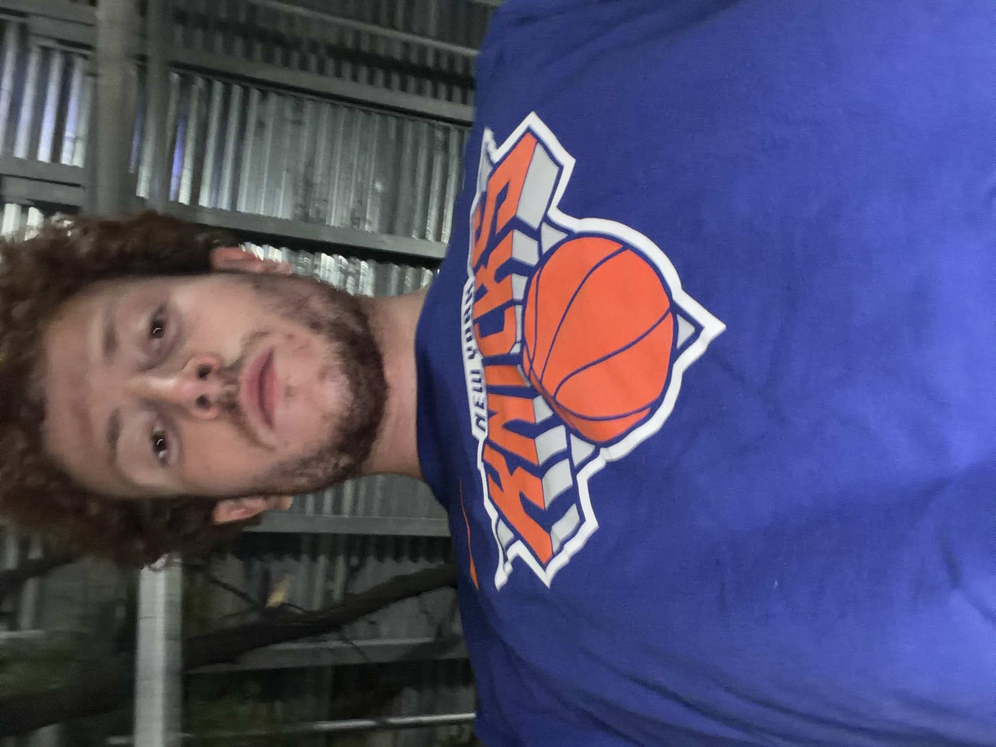 A person with curly brown hair and a beard is wearing a blue t-shirt with the New York Knicks logo on it. The background appears to be an urban outdoor setting, possibly a street or an area near a building with corrugated metal siding. The specific location in New York City cannot be determined from the image.