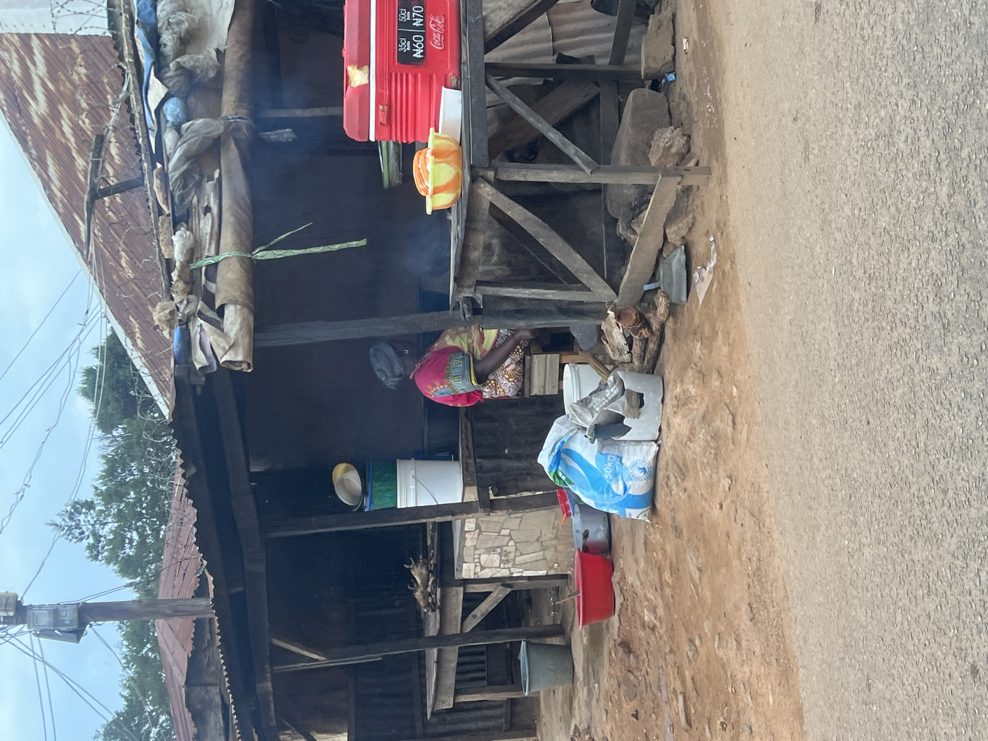 The image depicts an outdoor market stall in Jos, Nigeria. A woman is seated behind a wooden counter, engaged in food preparation. She is wearing a brightly colored patterned dress and headscarf. In front of her, a red cooler box with Coca-Cola branding sits on a table, next to a bowl containing orange slices. Various containers, buckets, and bags are scattered around the stall, including a prominent 50kg bag of what appears to be flour. In the background, a rustic structure with a corrugated iron roof is visible, along with trees and utility poles with overhead wires. The ground is a mix of dirt and debris. The overall scene suggests a busy marketplace environment.