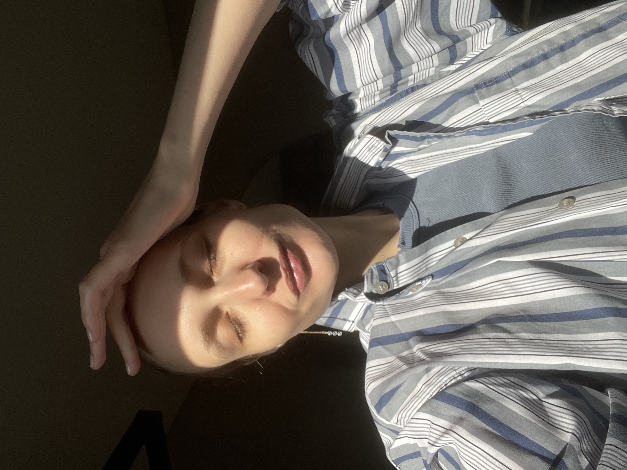 A person with light skin is lying down, their eyes closed, with their hand resting on their forehead. They are wearing a striped shirt with a button-down collar. The image is split into light and shadow, with the person's face brightly lit and the background dark. No information is available from the picture about the location of Подшивалово, Russia.