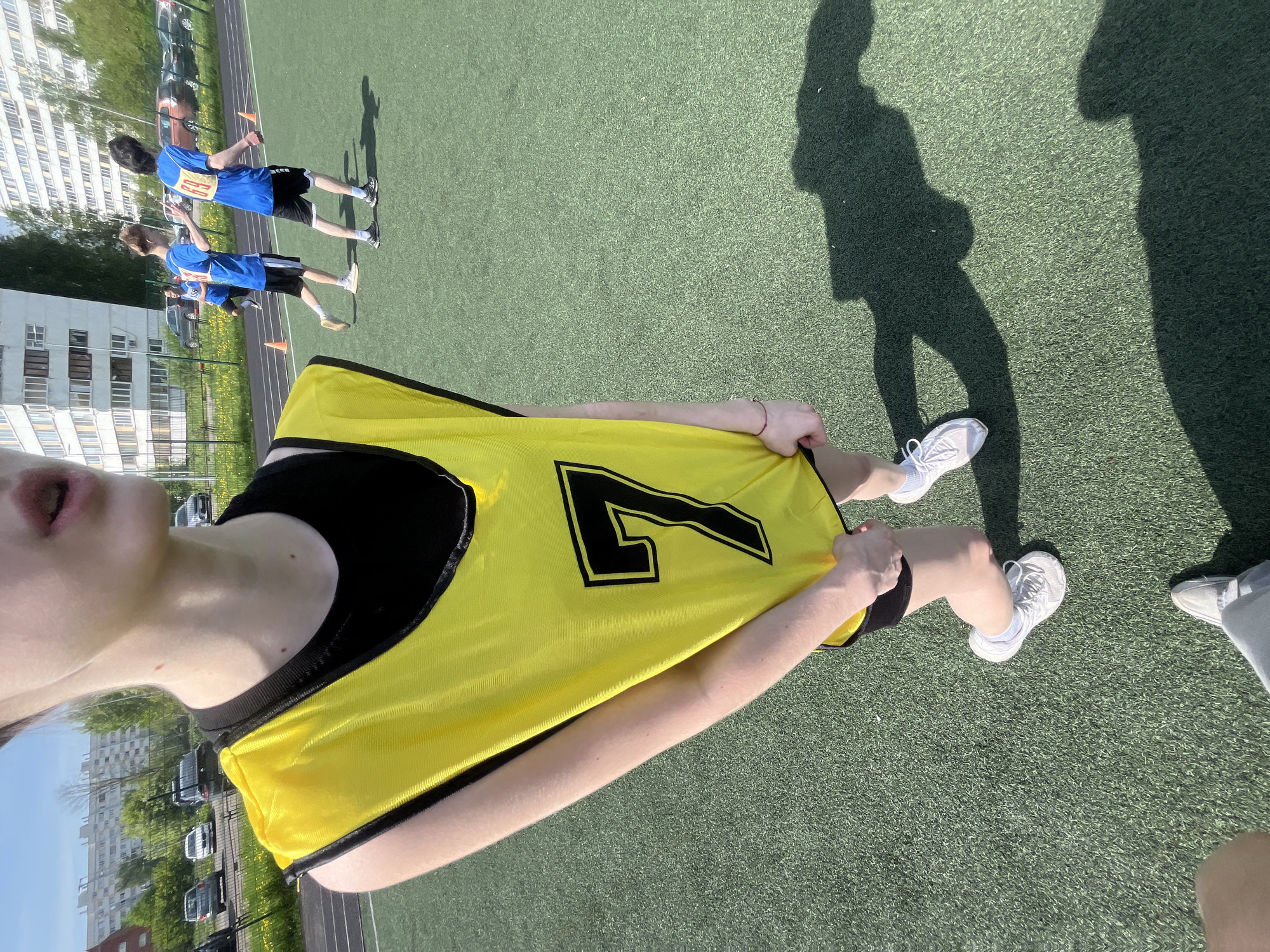 The image presents a youth sports event, likely taking place on an artificial turf field. A person wearing a yellow sports vest with a large black number "7" is in the foreground. Two other people in blue jerseys with numbers are doing sprints in the background. There's an apartment building in the background. Shadows of figures suggest additional people are present but not in direct view. The location appears to be Saint Petersburg, Russia, based on the architecture of the building in the background. The weather is sunny, as evidenced by the sharp shadows.