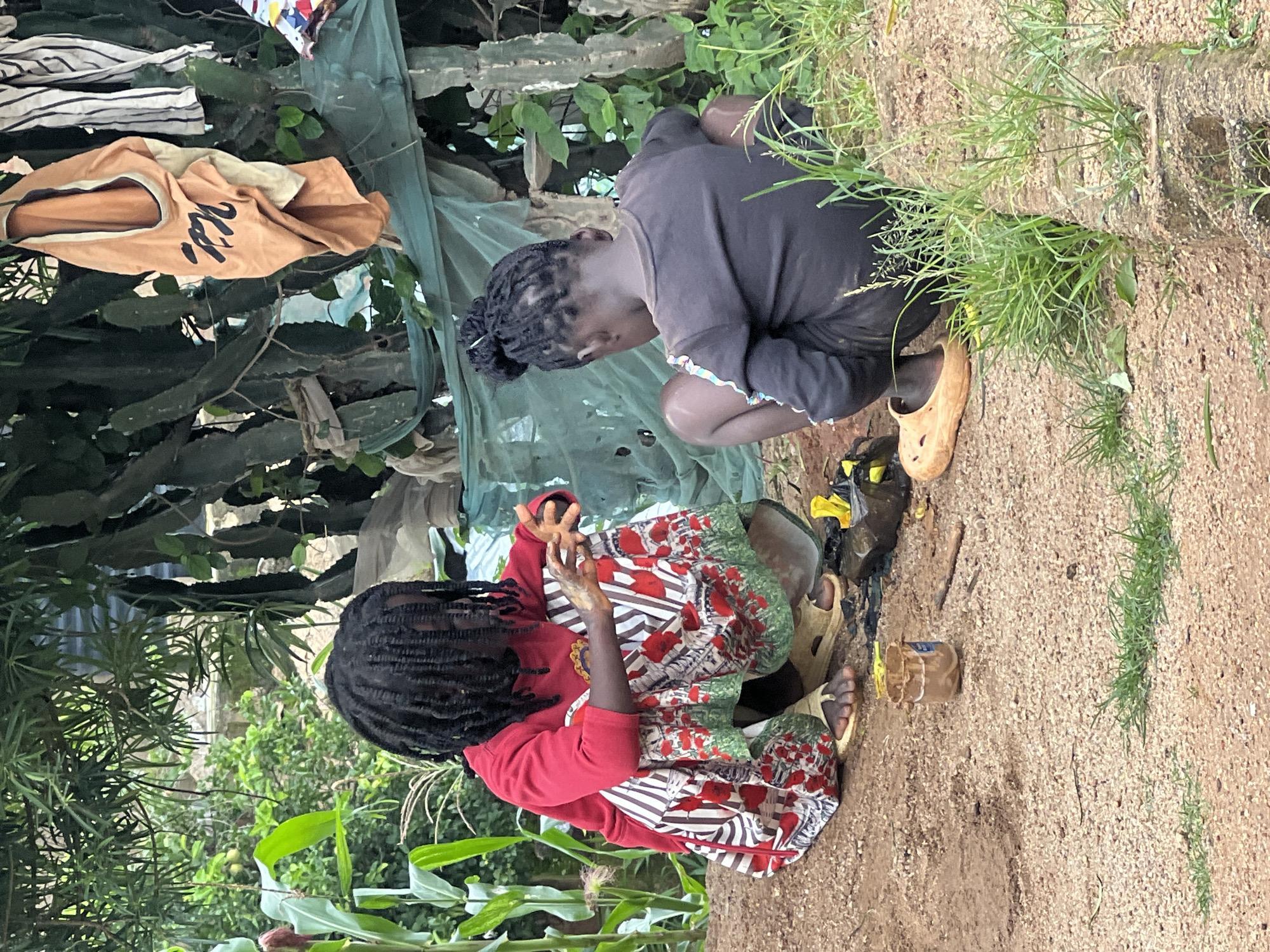 Two young children are crouching on a dirt ground, possibly in the city of Jos, Nigeria, engaged in an activity. The child on the left, wearing a red long-sleeved shirt and a patterned skirt with red flowers and green trim, is facing away from the camera and toward the child on the right. This child has dark, braided hair that reaches their shoulders and is wearing light-colored sandals. Their hands are held near a small, brown object on the ground, suggesting they are manipulating something.
The child on the right, wearing a dark grey long-sleeved shirt and dark shorts, is also facing away from the camera and toward the child on the left. This child has their dark hair tied up in a bun and is wearing light-colored, perforated clogs.
Between the children, on the ground, are various objects including a small brown container with green material sticking out, some sticks, and what appears to be a black bag.
The background is a mix of lush greenery, including corn stalks on the left, and a structure made of what looks like concrete blocks and possibly some salvaged materials covered with a green tarp or netting. In the upper right, clothes are hanging on a tree branch, including a bright orange garment with black writing. The overall environment appears to be an outdoor, semi-rural or informal urban setting.
