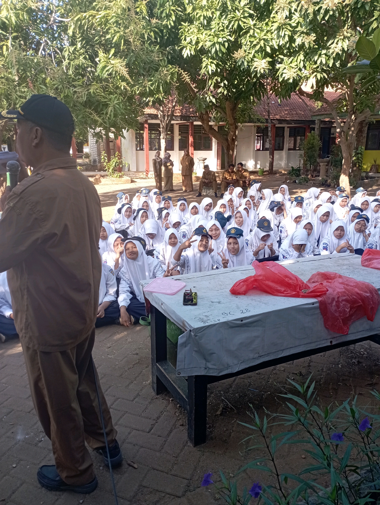 A man in a brown uniform and dark cap speaks into a microphone, facing away from the camera, at a school event. He stands in front of a large group of children, mostly girls wearing white hijabs and uniforms, seated on the ground. Behind the children, several adults in brown uniforms are visible. A white-covered table is in the foreground, displaying two red plastic bags and some paperwork. The scene appears to be outdoors, likely in the courtyard of a school in Banyuurip, Indonesia, with trees and buildings in the background. The lighting suggests it is daytime, possibly morning or afternoon.
