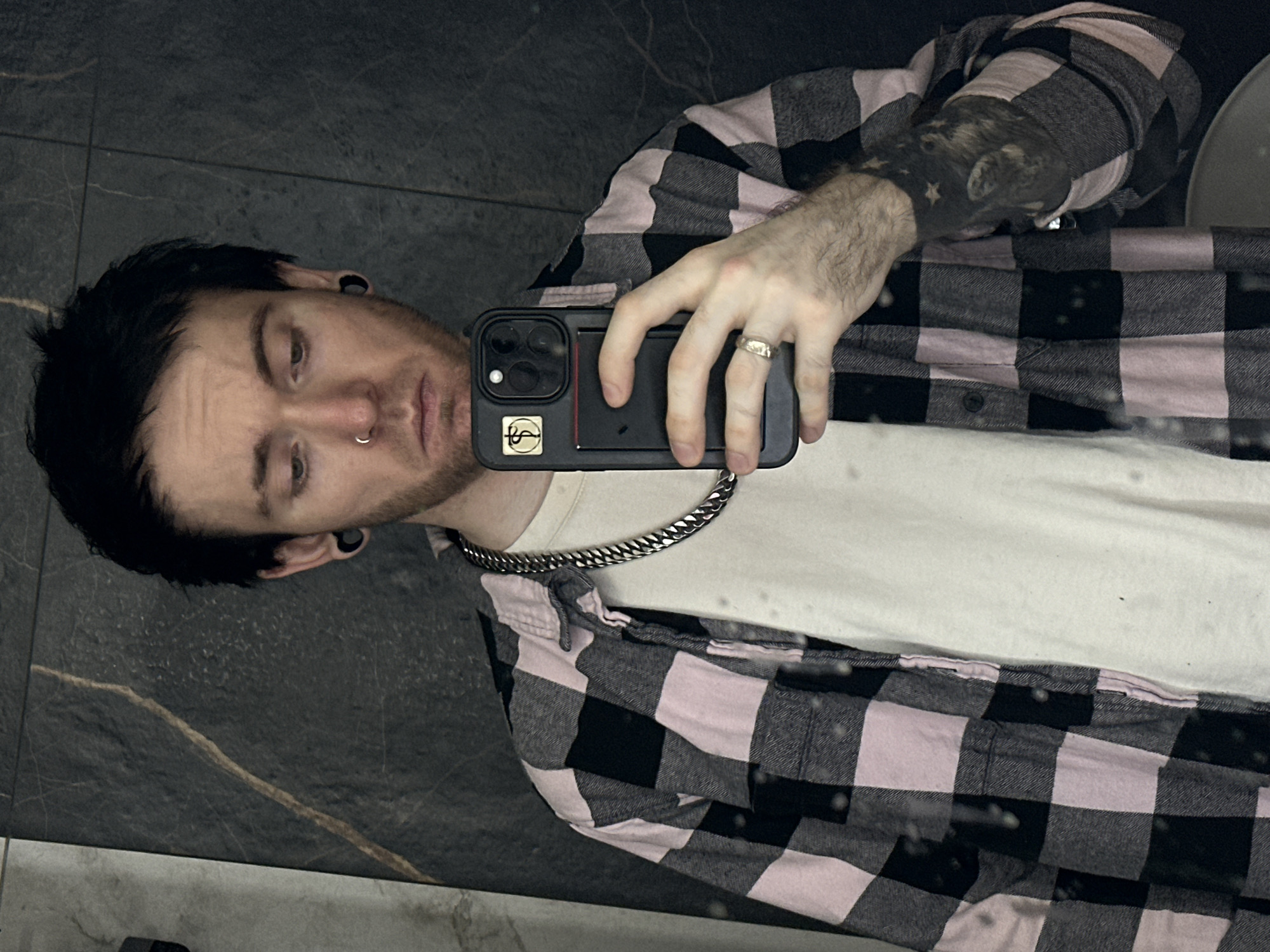 A man with dark hair, a nose ring, and stretched ear piercings is taking a selfie in what appears to be a bathroom. He is wearing a white t-shirt, a silver chain necklace, and a pink and black checkered flannel shirt. He has a tattoo on his left arm. He is holding a black smartphone with a design on the back. The background features dark, patterned tiles. There is no evidence to confirm the city of Khimki, Russia as the location.