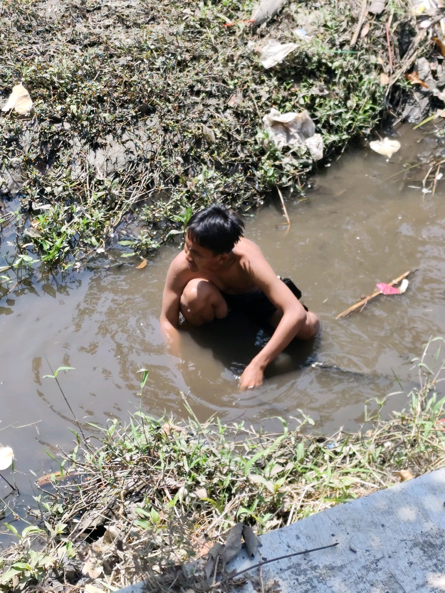 A young boy, with dark hair and a bare torso, is partially submerged in murky brown water. He is looking to his right, with his left arm bent and resting on his knee. He appears to be in a squatting position. The water is surrounded by muddy banks covered in sparse green vegetation and dry leaves. There is some debris, including plastic bags and sticks, floating in the water and on the banks. A concrete structure is visible in the foreground. The scene suggests an outdoor environment in an area that may be prone to flooding or has poor drainage, potentially in Bambanglipuro, Indonesia.