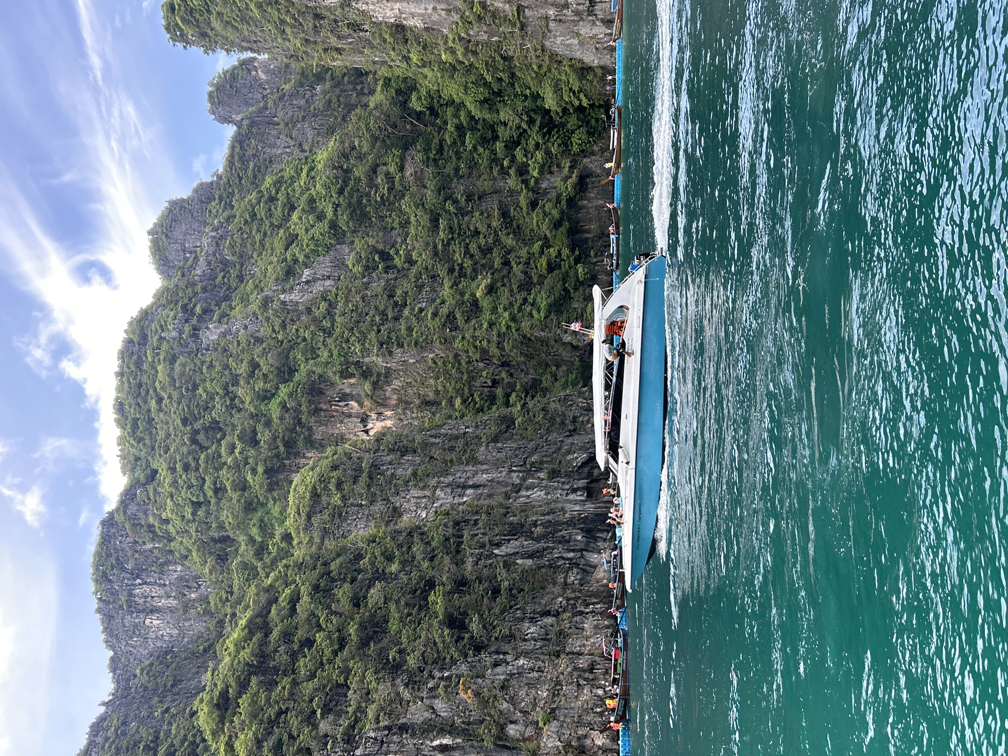 A speedboat with a blue hull and white cabin is traveling through emerald green water, creating a wake. The boat has people on board, some wearing orange life vests. In the background, steep, tree-covered limestone cliffs rise dramatically from the water, with a few other boats moored near the shore. The sky above is partly cloudy. This scene is characteristic of the island hopping tours and scenic bays around Ao Nang, Thailand.