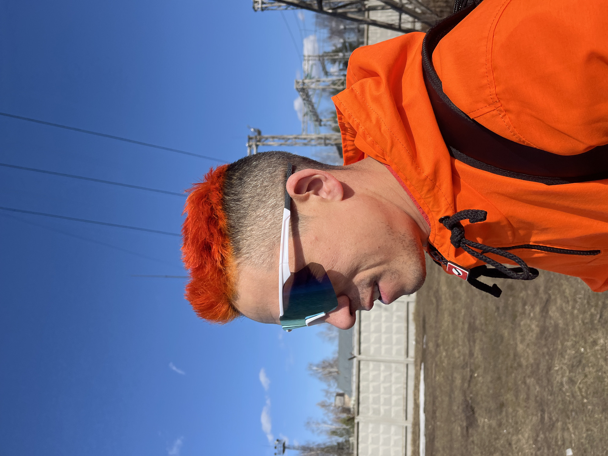 The image shows a person in Chaikovsky, Russia, outdoors on a sunny day. The person has short, gray-brown hair with a bright orange fringe. They are wearing white-framed sunglasses with reflective blue lenses and an orange hooded jacket with a black drawstring and zipper pull. The backdrop includes a pale blue sky with power lines and an electrical transmission tower. In the background, a concrete fence and trees are visible, along with patches of dry grass and snow on the ground. The photo is taken from a low angle, showing the person from the side.