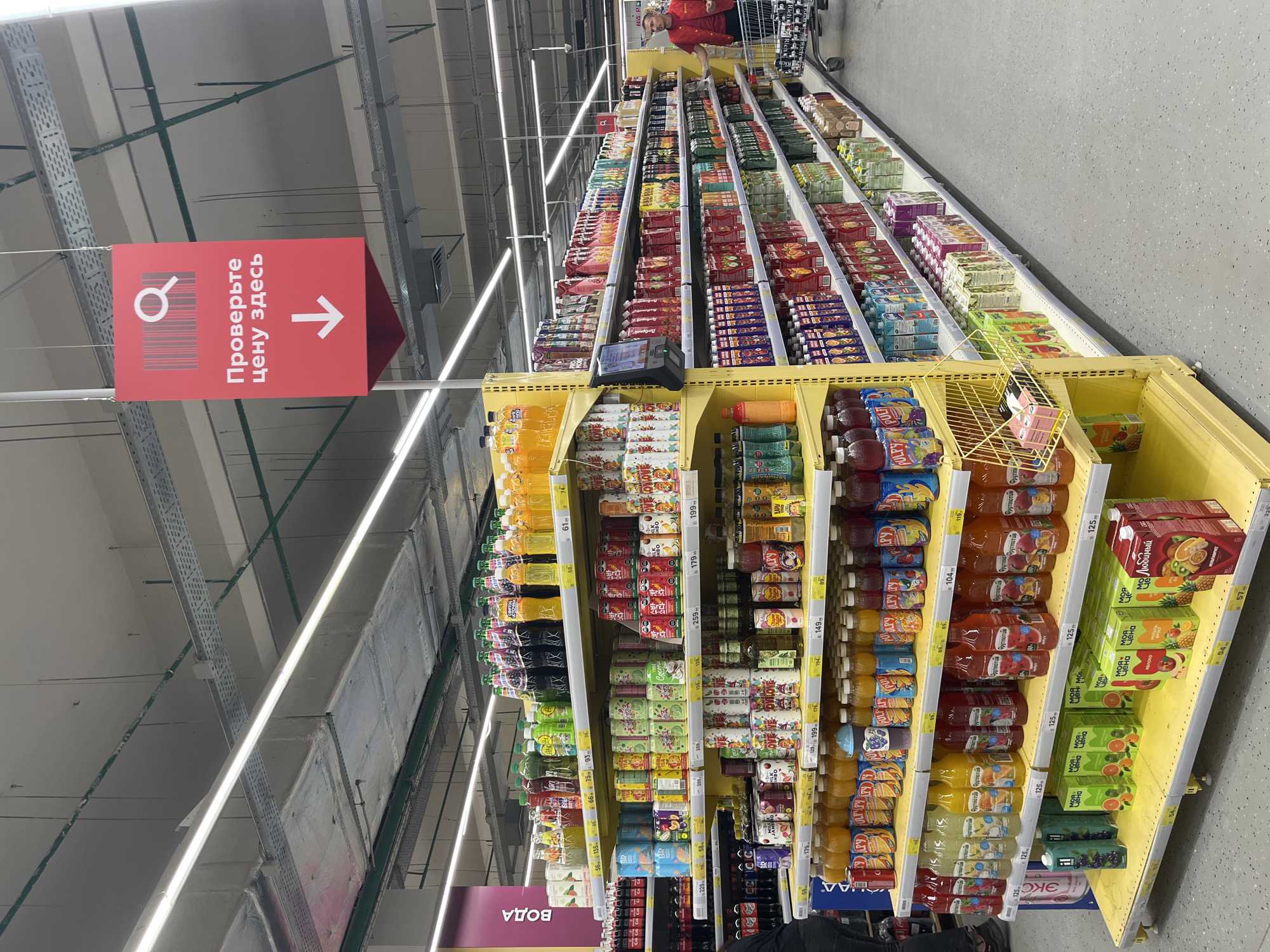 The image depicts the interior of a brightly lit supermarket, specifically a section dedicated to beverages. Rows of shelves are filled with various juices, sodas, and bottled water. Many products are packaged in colorful cartons and bottles, with prominent pricing displayed on yellow shelf tags. A prominent red sign with a barcode and a magnifying glass graphic hangs from the ceiling, with the Russian text "Проверьте цену здесь" (Check the price here) and a downward arrow, indicating a price checking station. A person wearing a red vest, likely a store employee, is visible in the background, pushing a shopping cart. The flooring is a speckled gray. The overall environment suggests a typical grocery store aisle in Volzhsky, Russia.