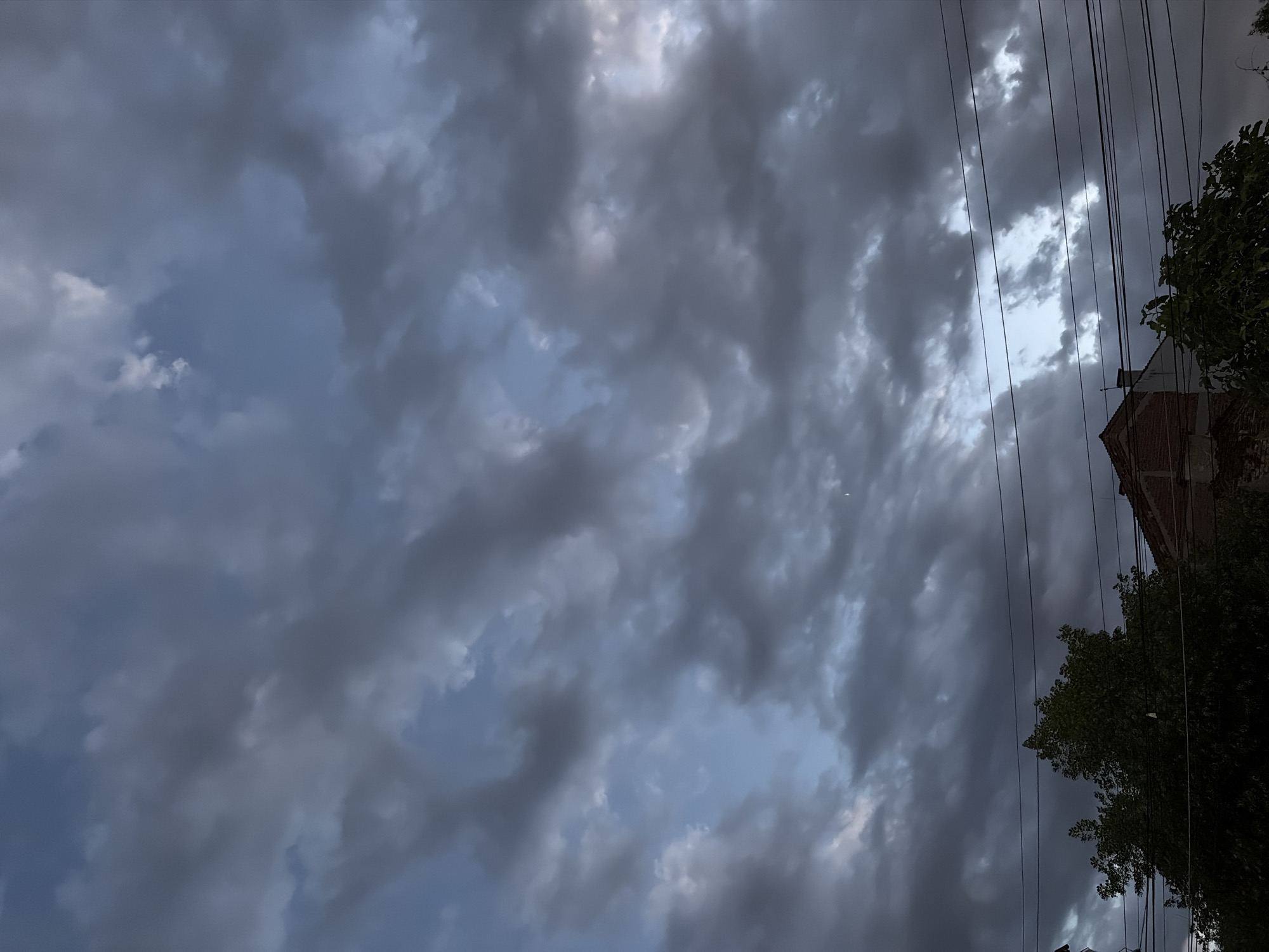 The image depicts a cloudy sky over a residential area. Dark, ominous clouds dominate the upper portion of the frame, with patches of lighter blue sky peeking through in some areas. The clouds are textured and appear to be moving, suggesting wind. In the lower half of the image, the tops of trees and the roof of a brick building are visible. Several power lines stretch horizontally across the scene, obscuring parts of the sky and the structures below. A faint crescent moon is visible in the upper right quadrant of the sky. The overall atmosphere is somber and hints at impending weather changes.