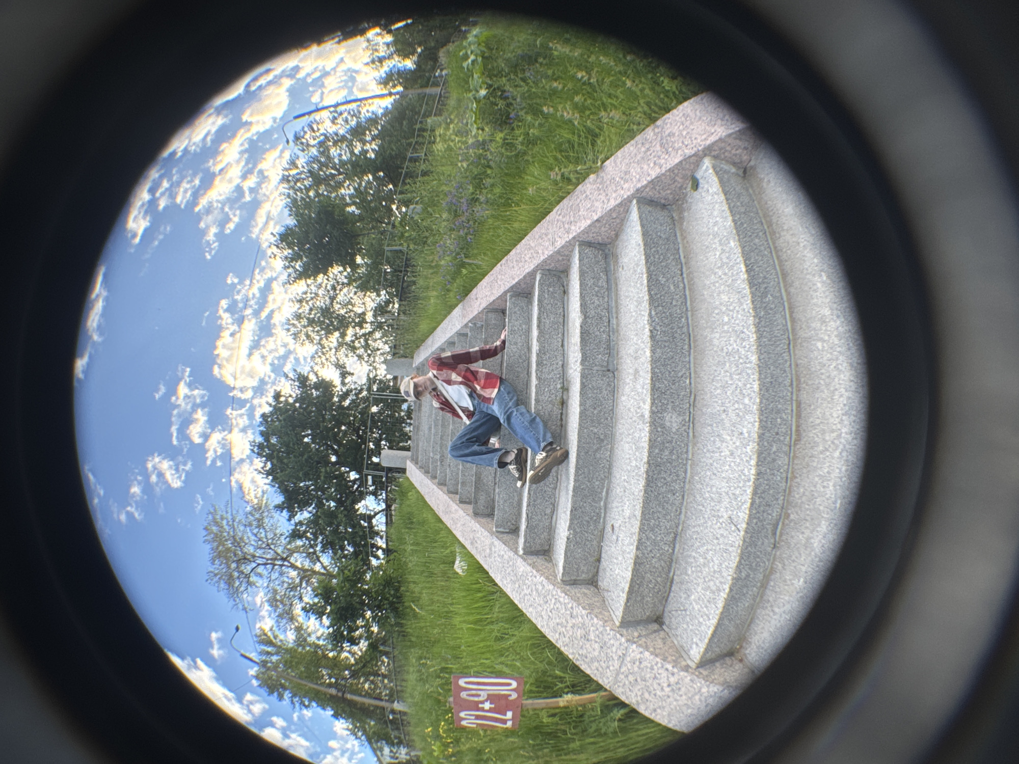 A young person wearing a red plaid shirt, blue jeans, a tan baseball cap, and brown shoes is sitting on a wide granite staircase. The person is looking towards the camera with their legs extended down the steps. To the left of the staircase, a red sign with white numbers "22+90" is planted in the grass. The staircase is surrounded by lush green grass and trees. The sky above is a bright blue with fluffy white clouds. The image has a fisheye lens effect, distorting the perspective. The scene appears to be a public park or a promenade.