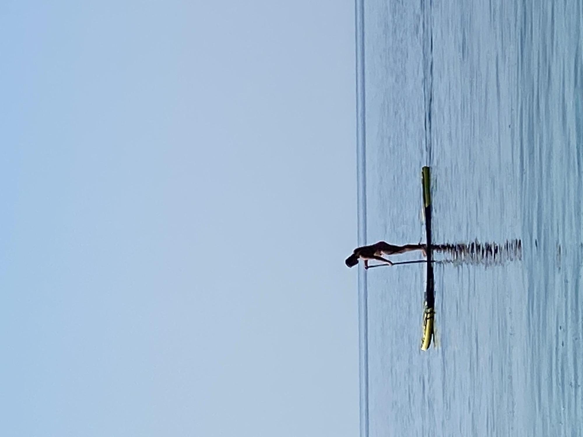 A solitary person stands on a yellow paddleboard in the calm, blue sea, holding a paddle. The person is facing left, with their head bowed. The paddleboard has some dark markings on its surface. The water is still, with a faint reflection of the paddleboard and person visible. The horizon is a thin, straight line separating the sea from a clear, light blue sky. The scene is tranquil and minimalist, with no other objects or people visible in the frame. This scene is located near the city of Prijevor, Montenegro.
