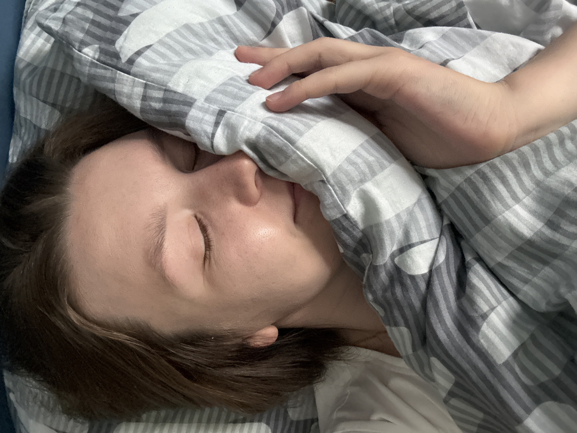 A close-up overhead shot shows a person sleeping. Their eyes are closed, and their face is partially obscured by a grey and white checkered comforter. Their right hand rests on the comforter near their face. The person is lying on a bed, with a blue surface visible at the top of the frame. The background suggests an indoor setting in Izhevsk, Russia.