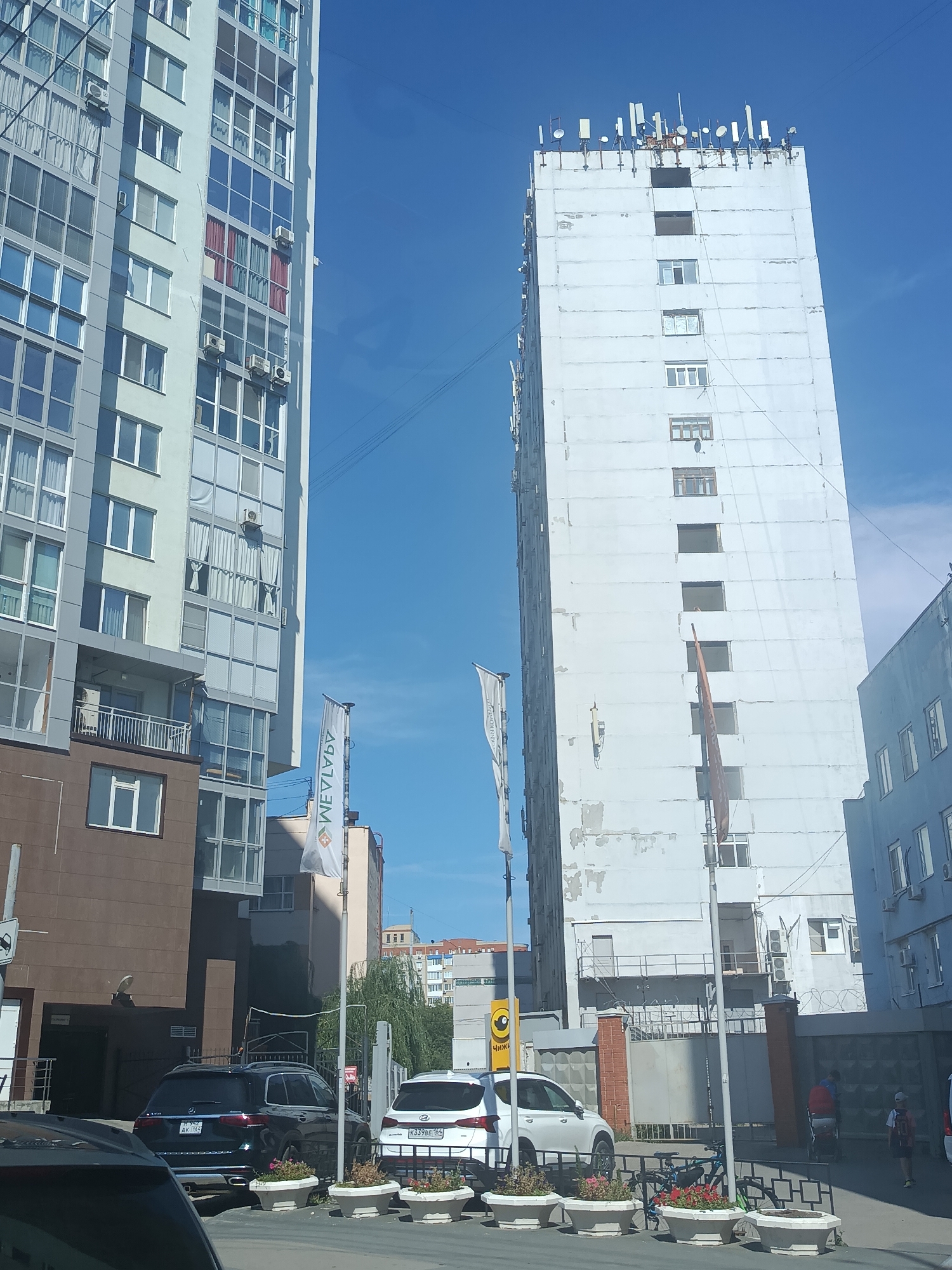 The image captures a street view in Saratov, Russia, dominated by two tall buildings. The building on the left is modern, with a light-colored facade and numerous windows, some featuring red curtains. Air conditioning units are visible on its exterior. The building on the right is older, with a white, weathered facade and a stark appearance. Its rooftop is densely populated with various antennas and communication equipment. A tall, dark structure, possibly a flagpole, is situated near the front of this building.
In the foreground, several cars are parked. A dark SUV, a white SUV, and a portion of another vehicle are visible. The dark SUV has a license plate starting with "M 742 AK". The white SUV has a license plate "K339BE 164". White planters with flowers are lined up along the sidewalk in front of the parked cars.
Two flags are positioned near the modern building. One is a white flag with a logo and text that appears to be "MEVLIA". Another flag is partially visible to its right. A street sign is mounted on a pole to the far left of the image. In the background, a glimpse of other buildings and trees can be seen under a clear blue sky. The scene depicts a typical urban environment with residential buildings and infrastructure.