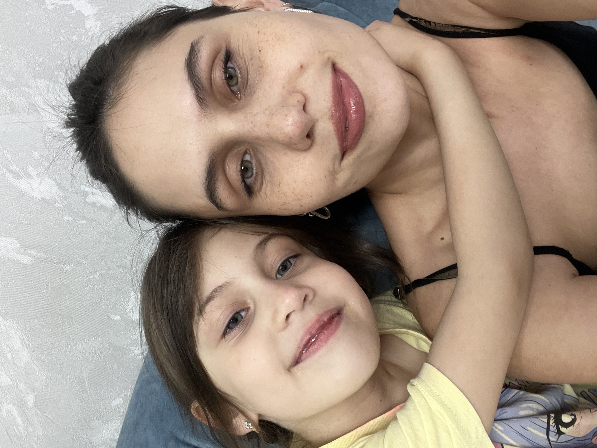 This intimate portrait captures two females, likely a mother and her young daughter, sharing a tender moment in what appears to be a home in Izhevsk, Russia. They are lying down closely together, with the adult woman's arm affectionately wrapped around the child.
The adult, positioned above and slightly behind the child, has dark hair, striking green eyes, and numerous freckles across her face. She wears subtle eye makeup, a rosy lipstick, and small silver earrings. Her attire includes a black strap or top. She looks directly at the camera with a gentle, content expression.
The young girl has light brown hair, soft blue-grey eyes, and a sweet, demure smile. She is wearing a bright yellow top and small, possibly floral, earrings. Like the adult, she is looking towards the camera, conveying a sense of calm and happiness.
The background reveals a textured light-colored wall on the left side of the adult's head and a blue soft surface, possibly a pillow or sofa, beneath them. A colorful patterned fabric, potentially a bedsheet featuring a cartoon-like eye and other elements, is visible under the child's right arm. The scene is brightly lit, suggesting an indoor setting. The overall mood of the image is one of warmth, affection, and familial closeness. There is no visible text.