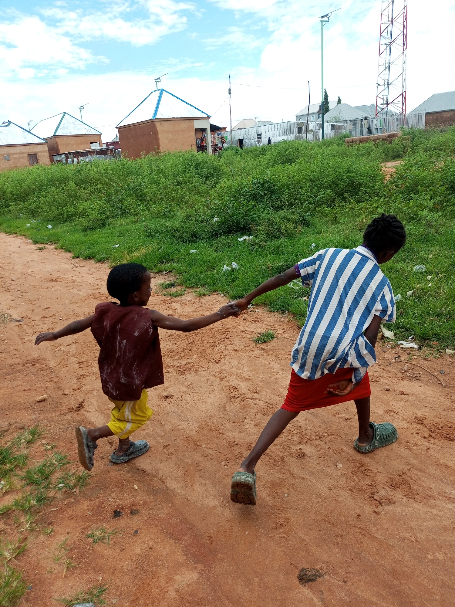 Two young children are walking hand-in-hand on a dirt road in Tempure, Nigeria. The child on the left wears a maroon sleeveless garment over yellow shorts and dark clogs. The child on the right wears a blue and white striped long-sleeved shirt, a red skirt, and green clogs. They are walking away from the camera, towards a backdrop of green vegetation and buildings with brown walls and white roofs, one of which has a blue accent on its roof. In the distance, a tall radio tower with red and white sections is visible against a cloudy sky. The ground is composed of reddish-brown dirt with patches of sparse green grass.