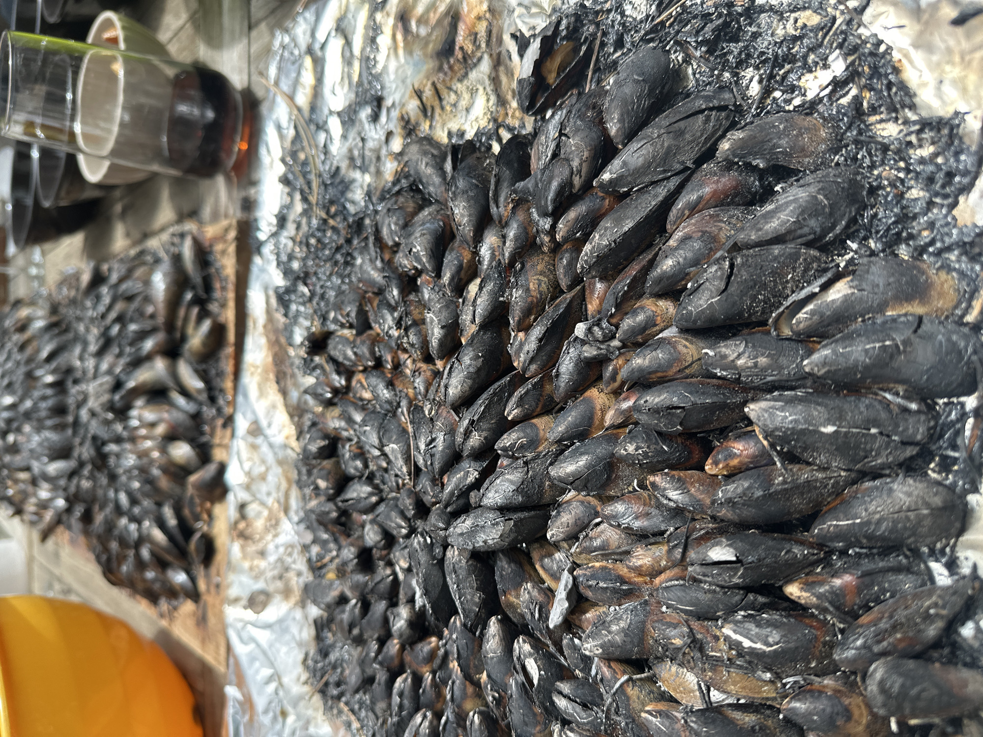 The image shows a close-up of cooked mussels, likely prepared outdoors, resting on a surface covered in aluminum foil. The mussels appear heavily charred, with a dark, almost burnt appearance. A smaller pile of mussels is visible in the background. Some glasses are partially visible in the upper-left corner of the frame. While the location provided is La Couarde-sur-Mer, France, there is no information in the image itself to confirm that this is where the photo was taken.