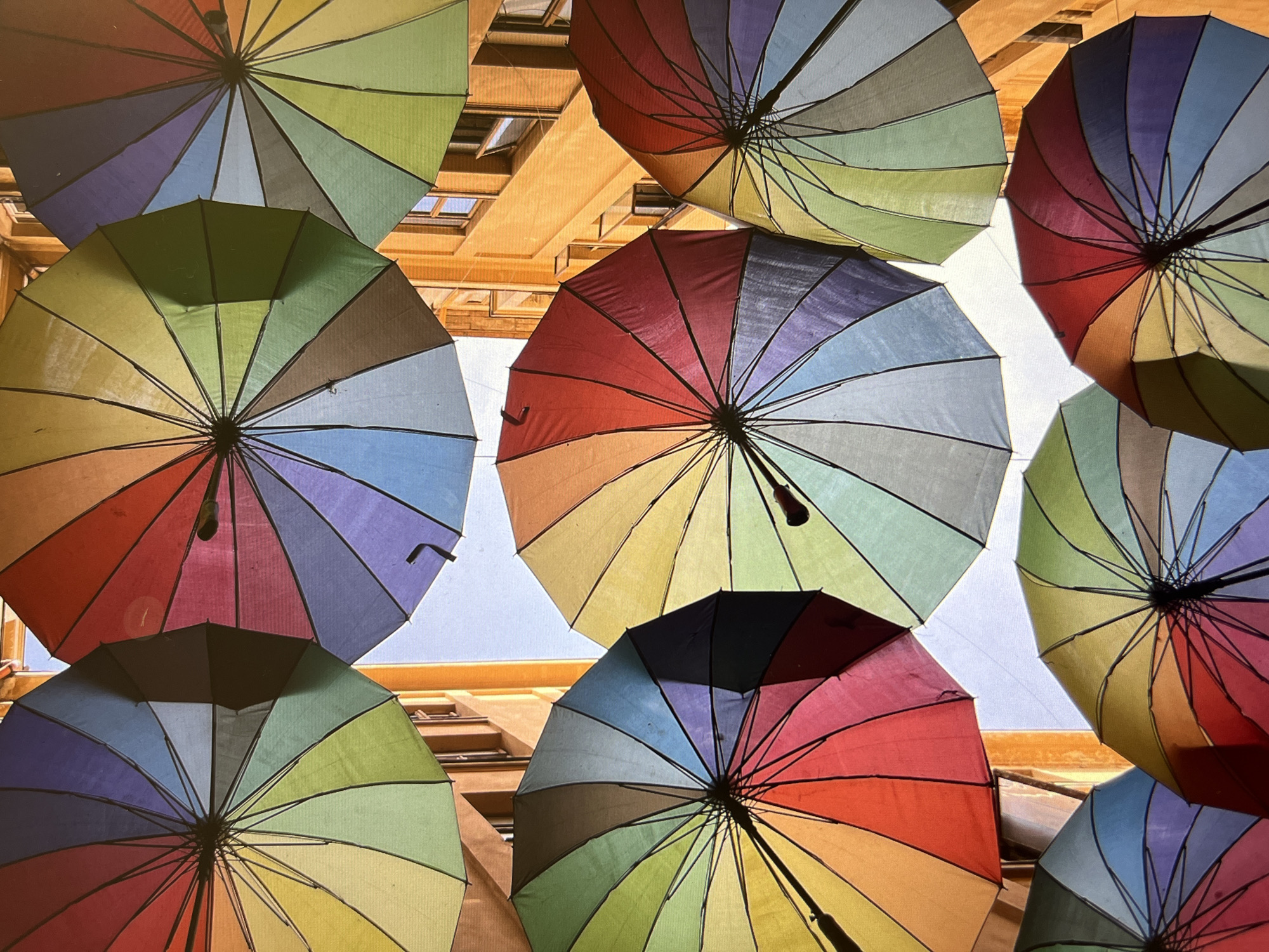Looking upwards, the image captures a vibrant display of open, multicolored umbrellas suspended in the air, possibly as a street decoration. The umbrellas are arranged closely together, creating a dense canopy of colors against the backdrop of buildings and the sky. The buildings appear to have a beige or light brown facade, with visible windows and architectural details. The sky is visible in small patches between the umbrellas, suggesting a bright, daytime setting.