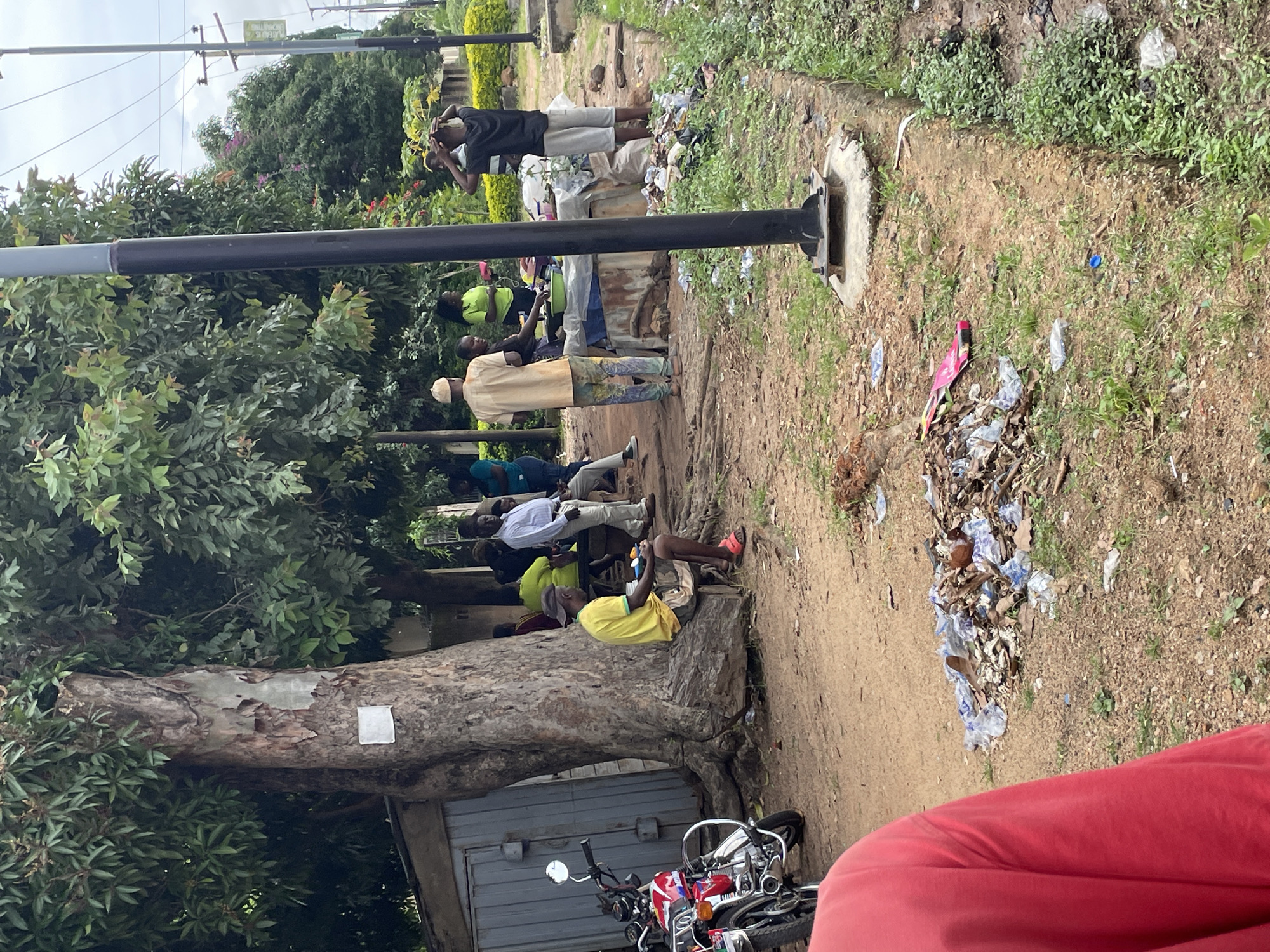 In Jos, Nigeria, under the shade of a large tree with exposed roots, a group of individuals are gathered. A man in a yellow shirt and shorts sits on a log, holding a small object. Nearby, a red motorcycle is parked. In the background, several people are standing and interacting around a makeshift table covered with a blue tarp. A black lamppost stands prominently in the foreground. The ground is littered with debris, including plastic bottles and dried leaves. In the distance, power lines and vegetation are visible, suggesting an outdoor urban setting.
