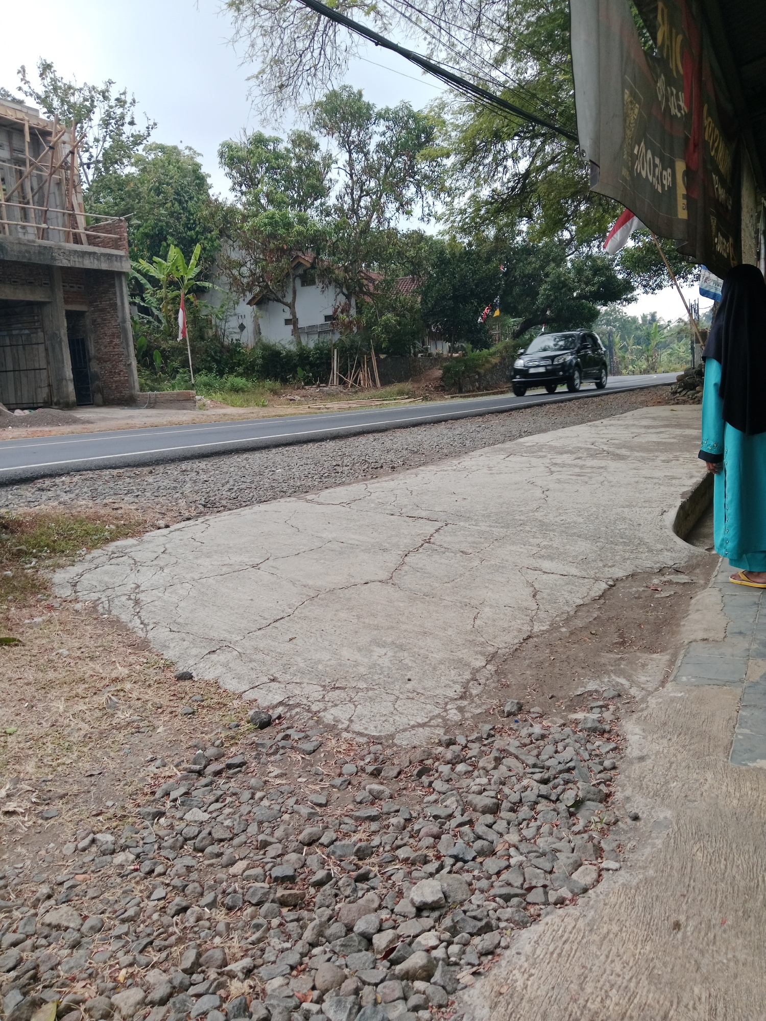 The image depicts a street scene in Bojongmengger, Indonesia. In the foreground, a cracked concrete pathway runs along the right side of the frame, bordered by a strip of gravel and dry grass on the left. A dark-colored SUV is driving on the asphalt road in the middle ground, moving away from the viewer. To the left of the road, a building under construction with exposed wooden scaffolding is visible, along with a small white house surrounded by lush green trees and vegetation. A red and white Indonesian flag is planted near the construction site. On the right side of the image, a person wearing a black hijab and a turquoise dress is standing on the concrete pathway, facing away from the camera. A dark banner with text is hanging from a structure above and to the right of this person, partially obscuring the sky and trees in the background. The sky is overcast, suggesting a cloudy day.