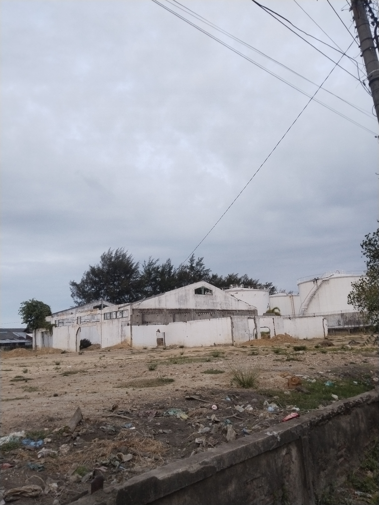 The image depicts a derelict industrial site in Lhokseumawe, Indonesia, under an overcast sky. Predominantly visible are decaying white-walled structures, including what appear to be storage tanks toward the rear. The foreground consists of a barren, dirt-covered lot with scattered trash and debris. A weathered concrete barrier partially obstructs the bottom of the view. Overhead electrical wires run diagonally across the scene. No people or moving subjects are visible. The overall environment suggests abandonment and disuse.