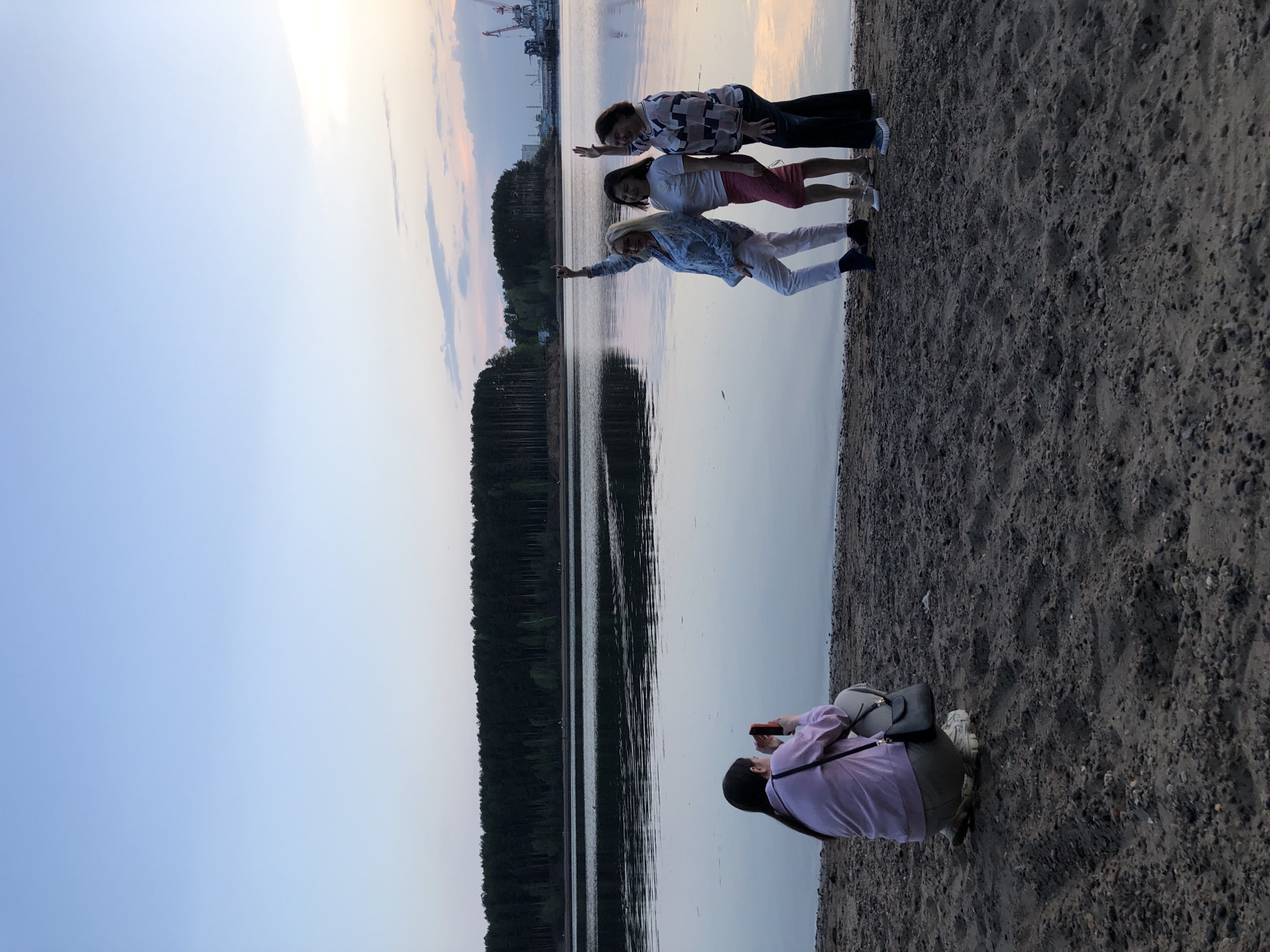 Here is a description of the image:
Three adult women are posing for a photograph on a sandy shore next to a body of water. They are arranged in a line, each resting their hands on the person in front of them, creating a human chain. The woman at the front is pointing her arm outwards.
Behind them is a lake, with a line of trees along the opposite shore. In the distance, there are industrial structures with cranes. The sky is a gradient of blue and light pink, suggesting either sunrise or sunset.
In the foreground, a person is crouched down, facing the group and taking their picture with a smartphone. They are wearing a pink shirt and carrying a purse or small bag.
The location is likely the city of Chaikovsky in Russia, indicated by the background landscape and industrial elements. The setting is a beach area adjacent to a lake or river.