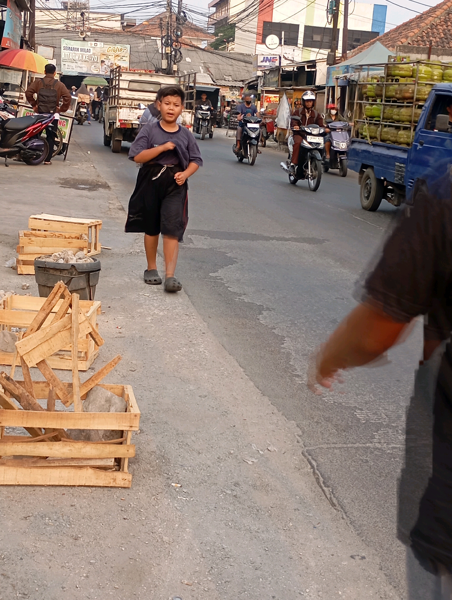 The image captures a street scene in Babelan, Indonesia. A young boy in a dark blue t-shirt and black shorts is walking on the right side of the road, facing towards the viewer. He is wearing grey Crocs. To his left, there are several wooden crates and a large black container filled with rocks. Further down the street, traffic is visible, including a blue truck loaded with green gas cylinders and several motorcycles carrying riders. The road is paved and appears to be somewhat worn. Buildings line the street, with some signs and power lines visible. The sky is overcast, suggesting it might be late afternoon or early evening.