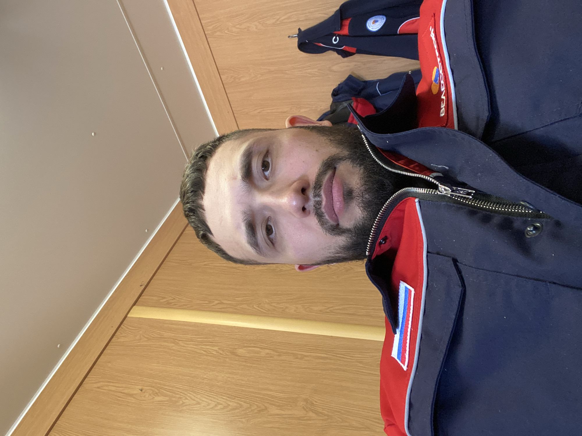 A man with dark hair and a beard is wearing a navy blue and red jacket with a Russian flag patch. He is positioned against a background of light brown wood paneling and a light-colored ceiling. There is another jacket hanging in the background. The location is Luzhitsy, Russia.