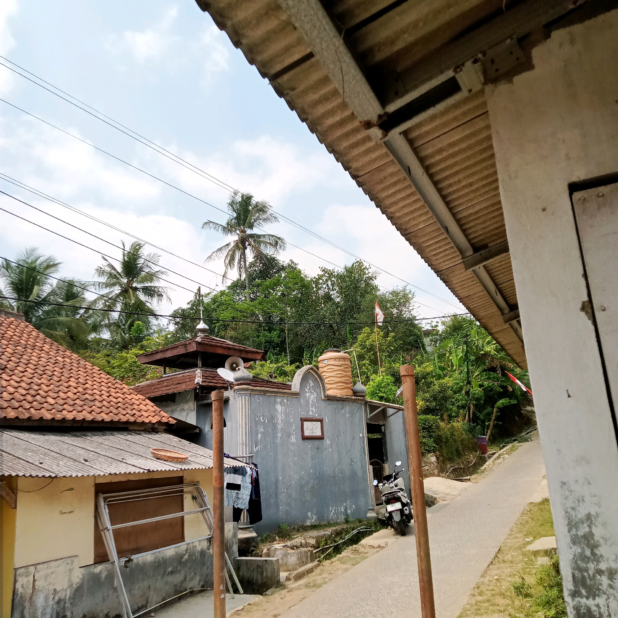 A narrow paved road winds through a densely vegetated, semi-rural area under a cloudy sky. On the left, a yellow building with a tiled roof is partially visible, adjacent to a corrugated metal roof structure with a drying rack. Further back on the left, a small building with a dome, likely a prayer house, is present. It features a satellite dish, loudspeakers, and a water tank on its roof. A motorcycle is parked on the right side of the road in front of a gray building with a framed artwork on its exterior wall. Lush green trees and palm trees dominate the background, with power lines crisscrossing the sky. An Indonesian flag is visible flying on a pole amidst the trees. The overall scene depicts a modest, tropical village setting.