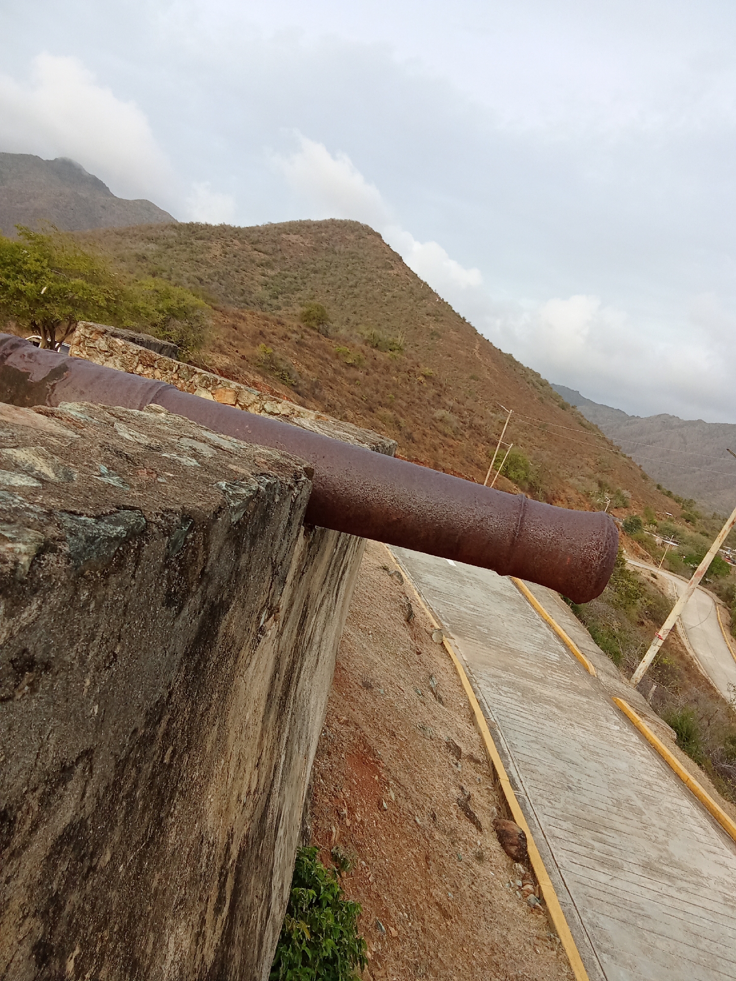 A slightly low-angle, medium shot shows an old, rusted cannon emerging from a stone fortification, likely situated on an elevated position in or near Santa Ana, Venezuela. The cannon extends horizontally over a concrete path or walkway with yellow painted edges. In the background, a rugged, mountainous landscape stretches towards a cloudy sky. A road winds its way through the hills, with utility poles visible alongside. The overall scene suggests a historic site or fort overlooking a mountainous region, possibly used for defense or as a landmark. The vegetation appears dry or sparse, typical of a semi-arid or seasonal climate.