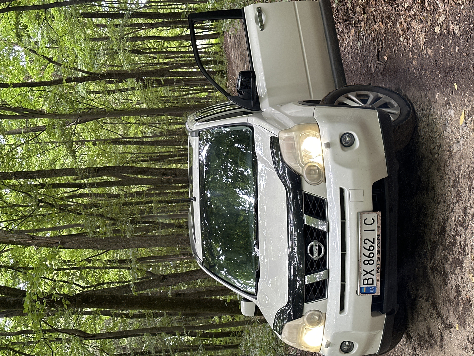 A white Nissan X-Trail SUV, with its driver's side door open, is parked on a dirt path in a forest. The vehicle's headlights are illuminated, and its license plate reads "BX 8662 IC" with a Ukrainian flag emblem. The car is positioned at an angle, facing slightly towards the viewer. The surrounding environment consists of a dense forest with numerous tall trees, their branches forming a canopy overhead. The ground is covered with dirt, fallen leaves, and some scattered stones. The scene appears to be in a natural, wooded area, consistent with a rural location in Kolubaivtsi, Ukraine. There are no people visible in the image.