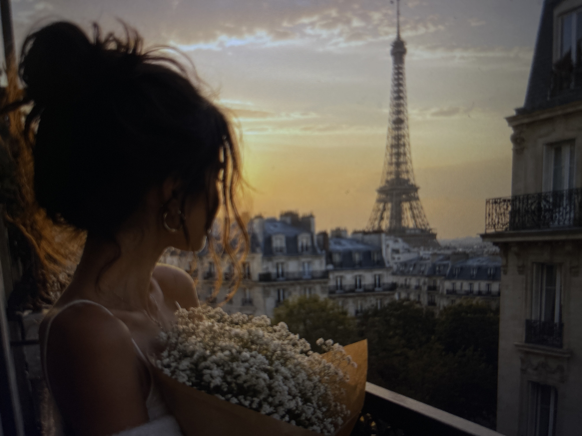 The image shows a woman on a balcony overlooking Paris. She is holding a bouquet of white flowers wrapped in brown paper. The Eiffel Tower is visible in the background, along with Parisian buildings. The sky is a gradient of orange and grey, suggesting either sunset or sunrise.