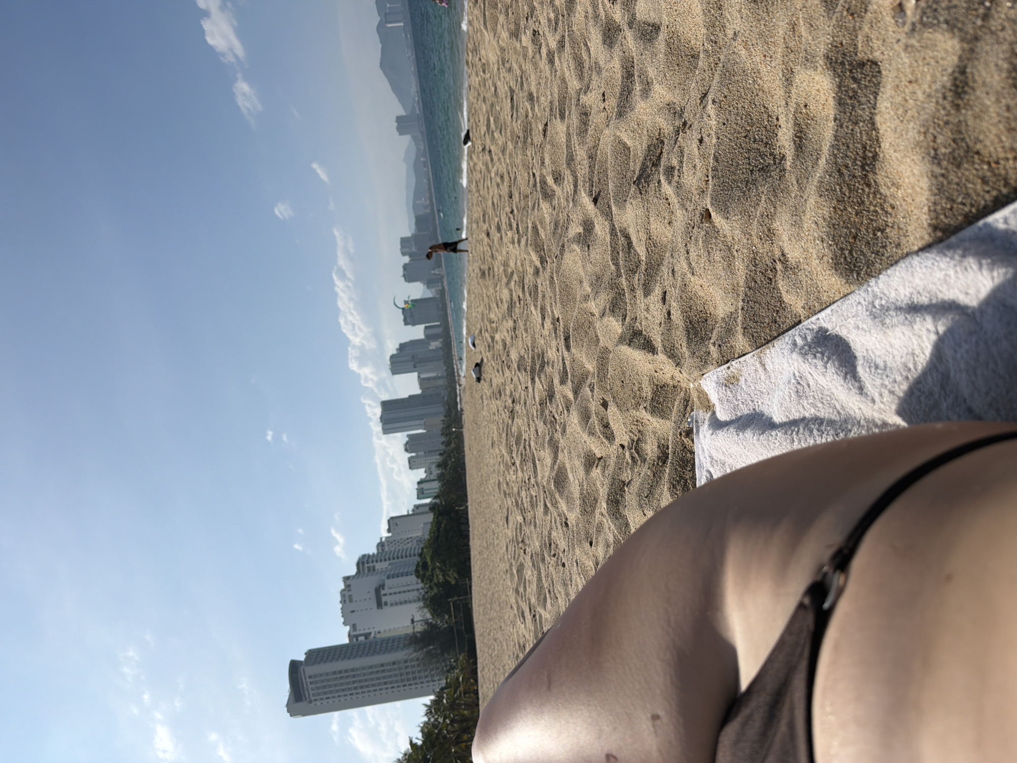 The image presents a first-person perspective from a beach in Nha Trang Ward, Vietnam. In the immediate foreground, a tanned leg and a dark bikini bottom with a silver ring detail are visible on a white towel, indicating a person lying on the sand. The midground features an expanse of light-colored sand with ripples and shadows, extending to the waterline. A man in dark swim shorts is walking left-to-right near the ocean's edge, where small waves are breaking. A colorful kite (green, yellow, blue) is visible in the clear blue sky above the water. In the background, a series of modern, light-colored high-rise buildings lines the coastline, partially obscured by a strip of green palm trees and foliage. Hazy blue mountains are visible in the far distance across the sea. The sky is clear blue with scattered white cumulus clouds, suggesting a bright, sunny day.