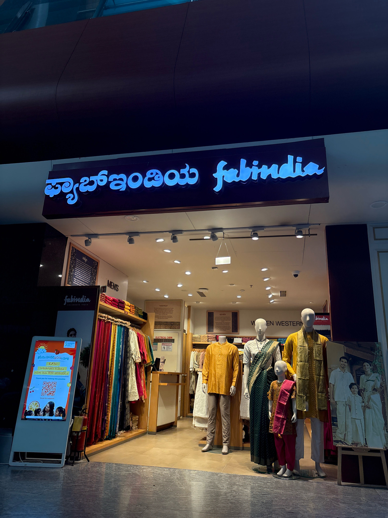 This image depicts the storefront of a Fabindia clothing store, likely located in Anneshvara, India. The prominent blue neon sign above the entrance reads "Fabindia" in both Kannada and English script. Inside the store, racks of colorful Indian garments are visible, along with mannequins dressed in traditional attire. A digital display screen to the left of the entrance shows a QR code and promotional text. The flooring is tiled, and the ceiling features recessed lighting. The overall impression is of a well-lit retail space showcasing Indian fashion.