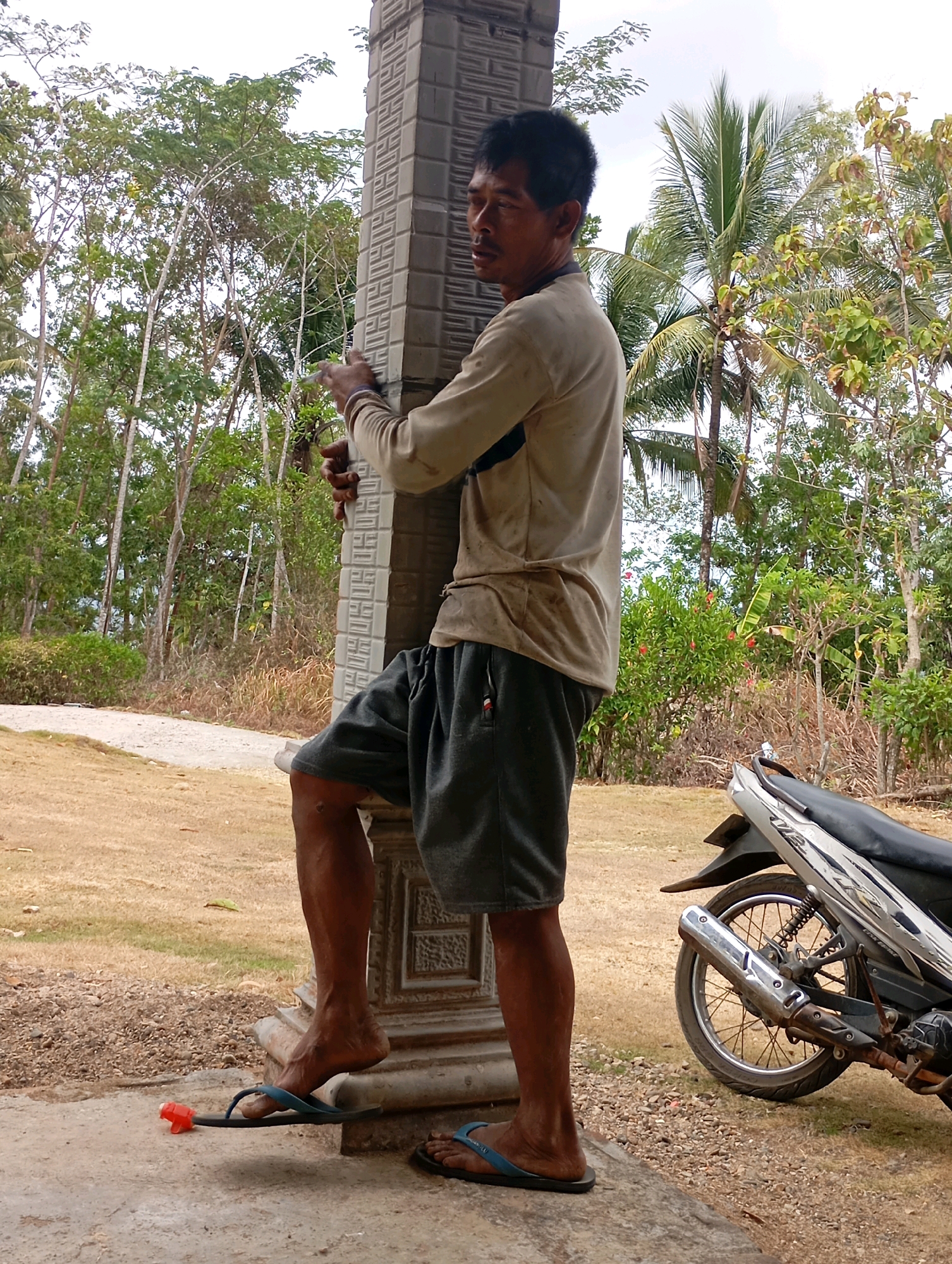 In Ketepung, Indonesia, a man with dark hair and a light-colored long-sleeved shirt and dark shorts stands with his right leg propped on a concrete pillar. His left foot rests on a dark sandal with a red object placed on it. A motorcycle is parked to the right of the man. The background consists of lush green trees and vegetation under a cloudy sky. The ground is covered in dry grass and dirt.