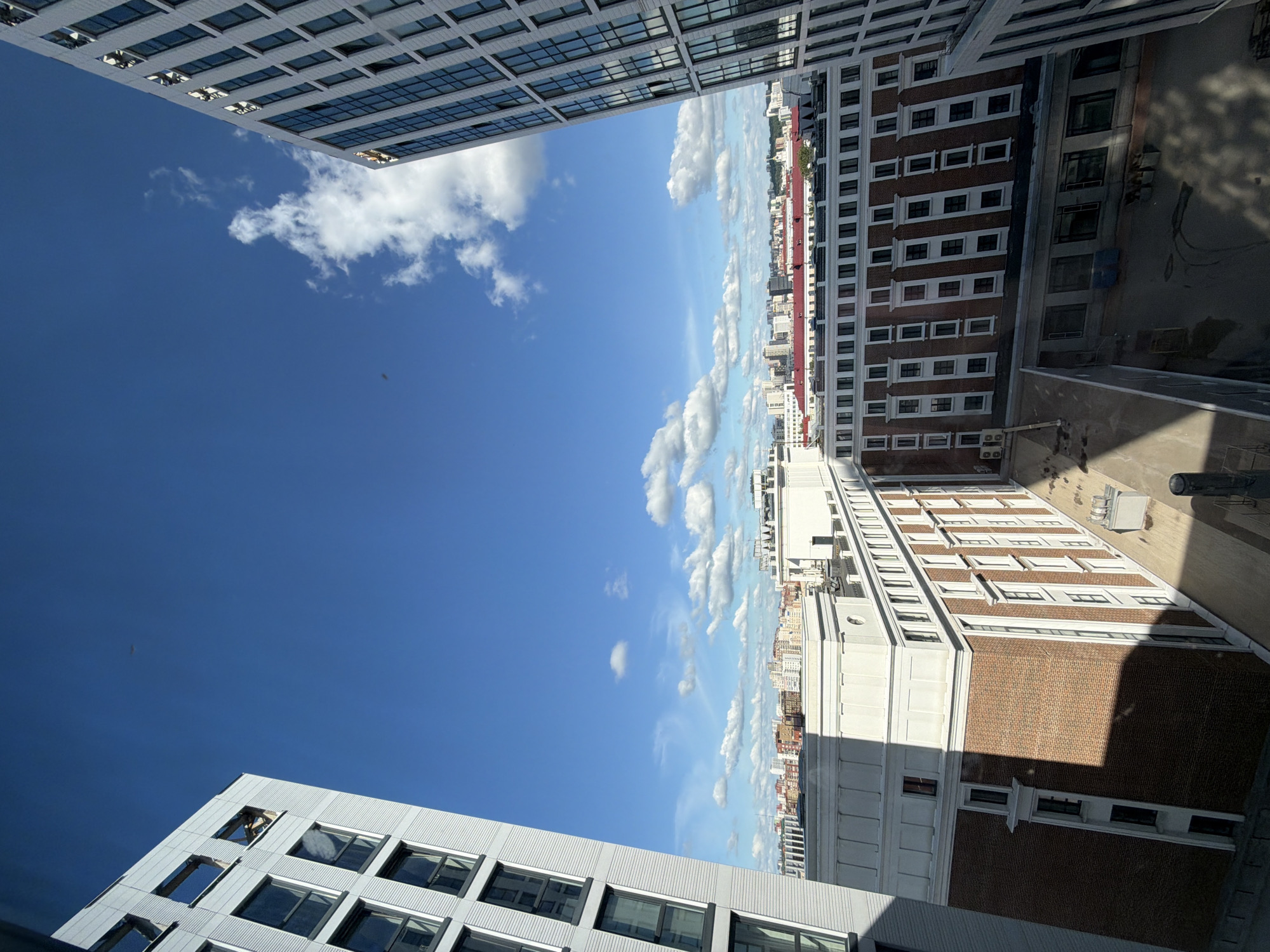 The image shows an exterior view of modern buildings against a bright blue sky with scattered white clouds. The perspective is from a high vantage point, looking down and across a cityscape. Several multi-story buildings are visible, characterized by repeating window patterns and varied facade materials, including brickwork and glass. One building in the foreground on the left has a light-colored exterior with rows of dark-framed windows. Adjacent to it, a taller building with a similar window design is partially visible. In the center and right of the frame, other buildings with brick facades and numerous windows are prominent. Some of these buildings appear to be older or have a more traditional architectural style compared to the modern structures. The scene suggests an urban environment, likely a city, given the density of buildings. Shadows are cast on some of the buildings, indicating the time of day is likely midday or afternoon. The weather appears clear and sunny. No people are visible in the image. There is no visible text. The location is Ufa, Russia.