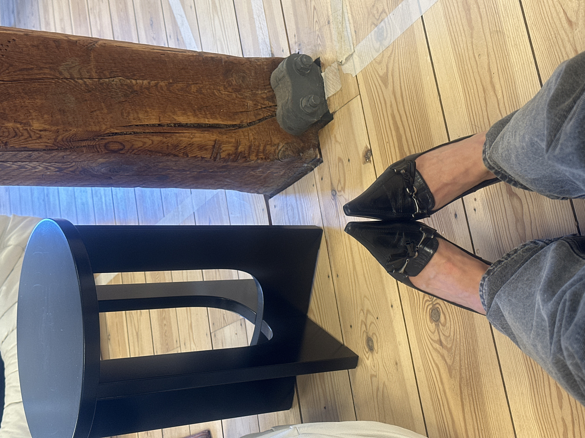 The image captures an indoor scene, likely in London, United Kingdom, showing a close-up, high-angle view of a person's legs and feet alongside architectural and furniture elements. The person is wearing light grey, perhaps faded blue, jeans rolled up at the ankles, paired with black pointed-toe heeled loafers featuring tassel and metal hardware details on the vamp.
The floor is composed of light-colored wooden planks with visible grain and knots. A prominent, thick, dark brown wooden beam with a rough, aged texture and several cracks extends horizontally across the upper left of the frame. This beam is secured to what appears to be a wall or support structure by a dark, heavy-duty metal bracket with two visible bolts. A piece of light-colored tape is visible near the floor-wall junction behind the bracket.
In the bottom left, a modern black side table or stool is partially visible. It features a circular top and a sculptural base with curved and angular elements, all in a matte black finish. The lighting suggests a bright indoor environment, possibly daytime. No specific activity is clearly depicted beyond the presence of the person's feet in a static pose. No text is visible in the image.