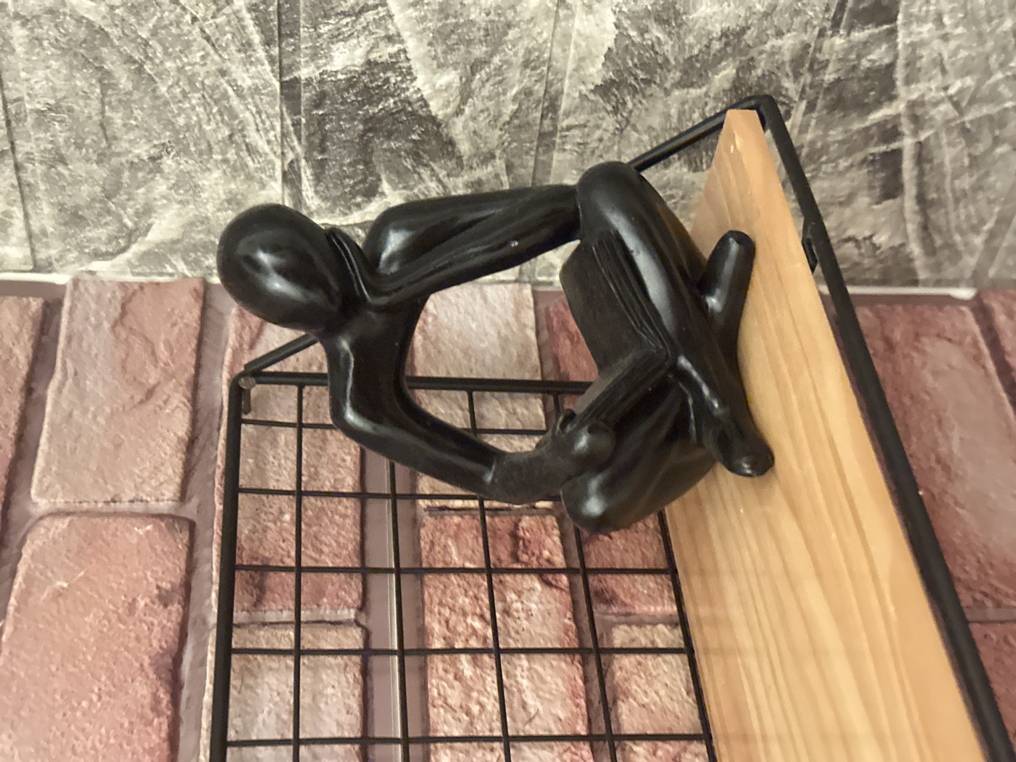 The image shows a black, abstract sculpture of a person in a seated, contemplative pose. The sculpture appears to be resting on a small, open-backed bookshelf or display stand. The shelf has a light wood surface and a black metal frame with a wire grid on the back.
The background consists of two different faux brick or stone wall coverings, one grey and one reddish-brown. No specific landmarks or architectural features indicate the location as London or any other city in the United Kingdom. There are no people in the image, and the focus is entirely on the sculpture and the shelf it rests on.