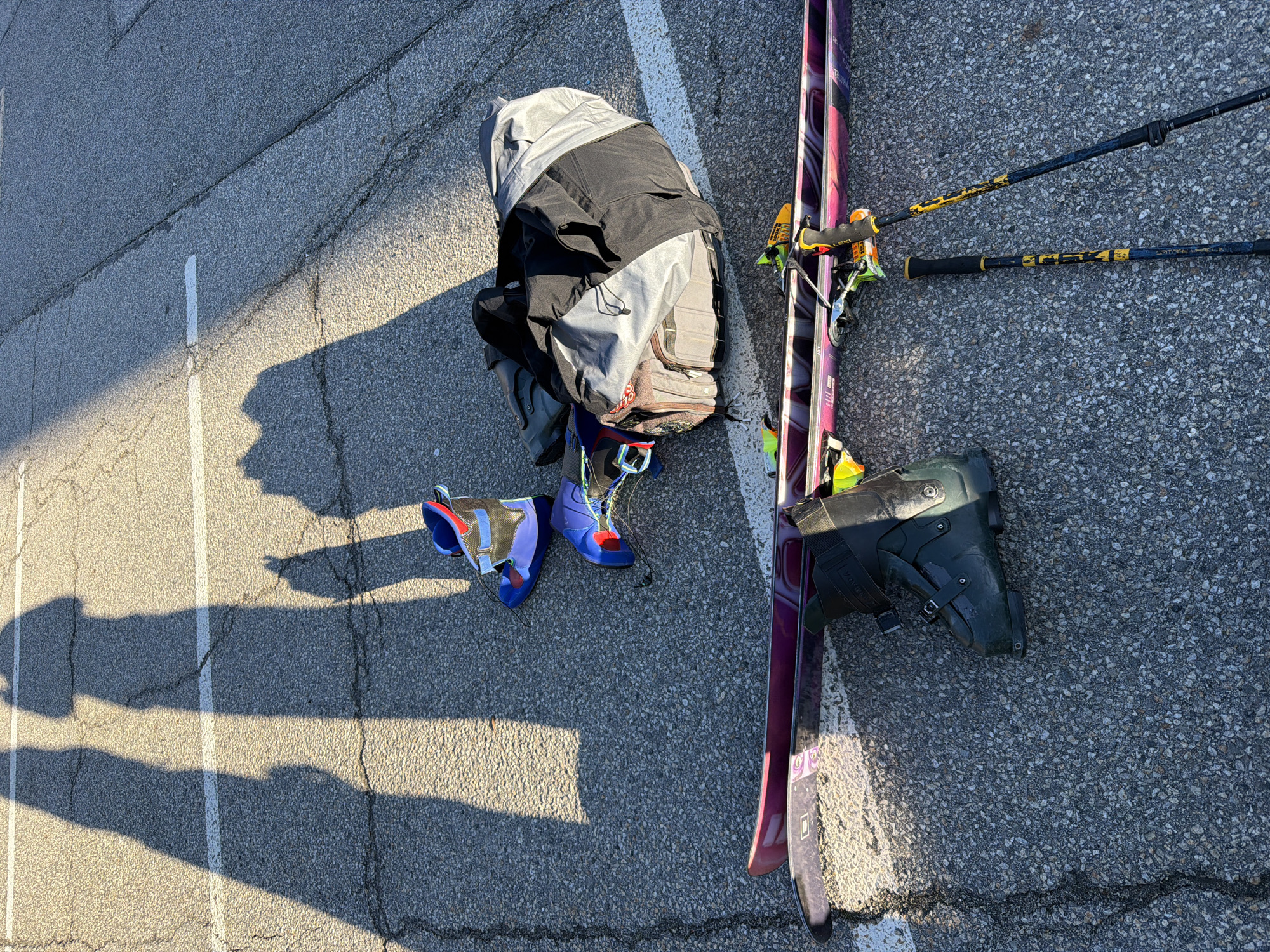 The image depicts ski gear laid out on an asphalt surface, likely a parking lot, as indicated by the white painted line. Gear includes: a partially open grey and black ski bag, a pair of purple and pink skis with yellow bindings and ski poles, and two pairs of ski boots (one pair is blue, purple, red and grey, and the other pair is dark green. The scene is bathed in strong sunlight, casting prominent shadows of people. The location is potentially Orelle, France, considering the presence of ski equipment, suggesting proximity to a ski resort.