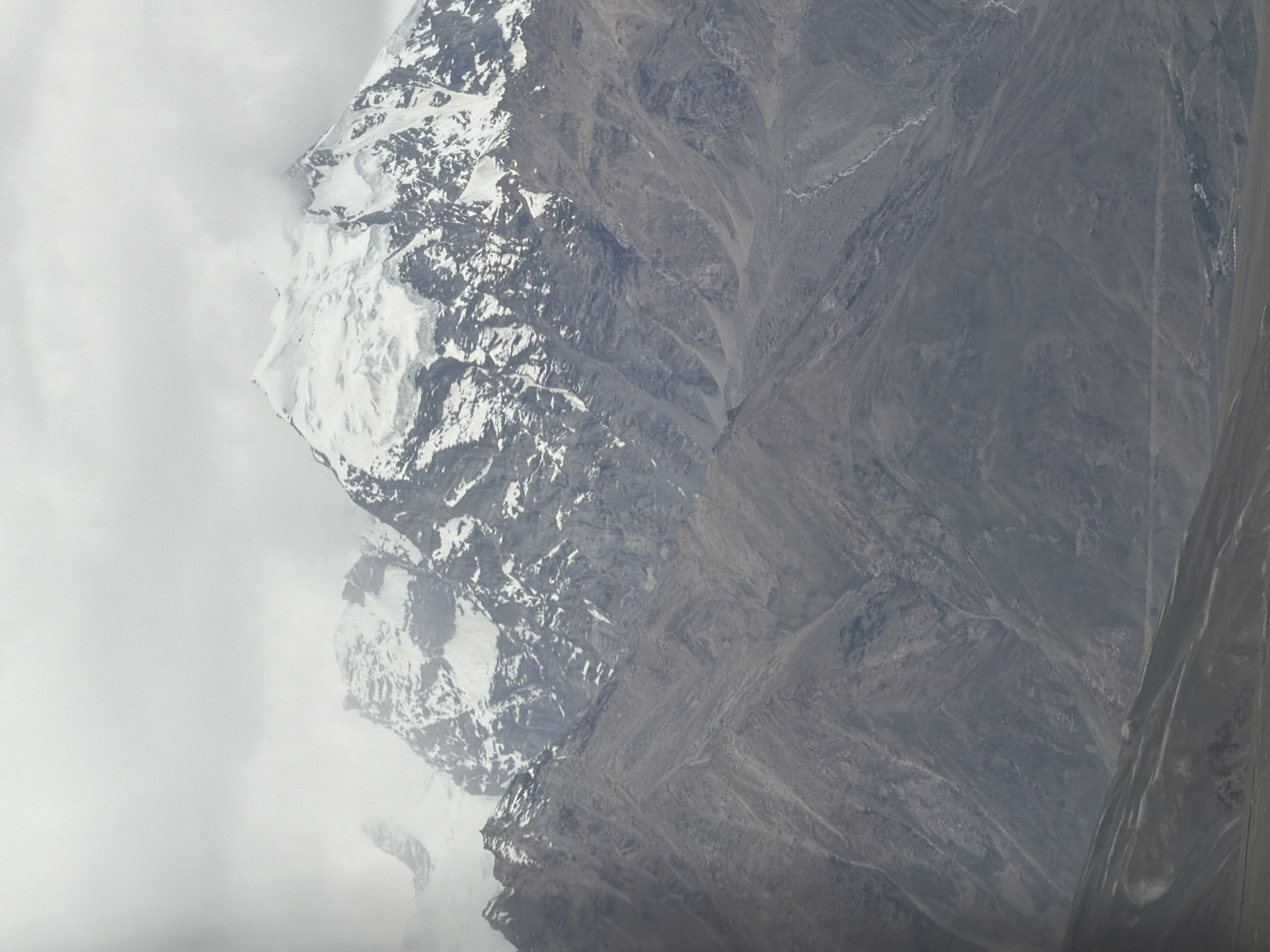 A vast mountain range dominates the frame, with snow-capped peaks piercing through a heavy, overcast sky. The lower slopes of the mountains are a muted gray, textured with rock and scree. A winding, lighter-colored path, possibly a riverbed or road, is visible on the right side of the mountains. In the distance, at the base of the mountains, a cluster of small buildings can be seen, suggesting a settlement. The foreground is a blur of muted, undulating terrain, hinting at foothills or a valley floor. The overall atmosphere is one of rugged, imposing natural beauty, with a stark contrast between the bright, snow-covered summits and the shadowed, rocky lower regions. The image appears to be taken from a distance, capturing the grandeur of the landscape. There are no people or animals visible in the scene. The location is identified as Stongde, India.