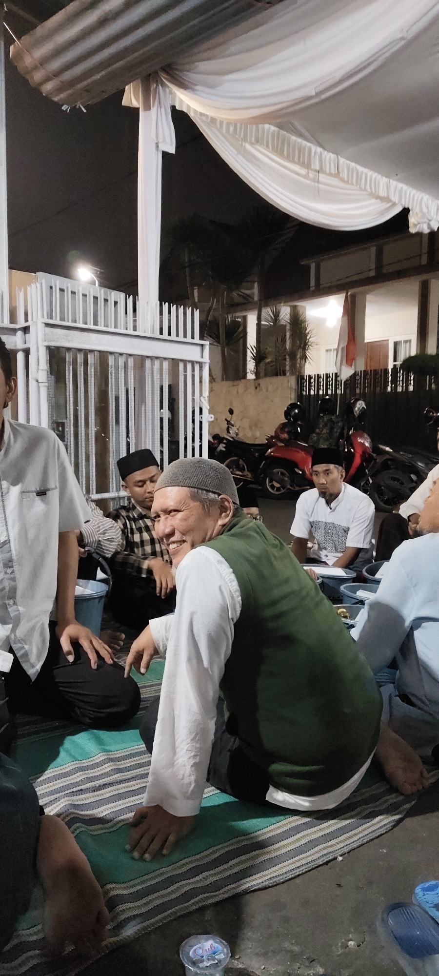 The image depicts a gathering of several men sitting on the ground, likely a street or an outdoor communal area, in Malang, Indonesia. A white tent structure is erected overhead, providing shade. In the background, a house with an Indonesian flag is visible, along with parked motorcycles, suggesting a residential or community event.
The men are dressed in traditional attire, including peci caps and long-sleeved shirts. One man in the foreground, wearing a green vest over a white shirt and a grey peci, is looking at the camera and smiling broadly, his hand gesturing forward. Another man in the background, also wearing a peci and a checkered shirt, is looking down. A third man, seated further back, is wearing a white shirt with a patterned detail on the chest and a black peci. Several plastic containers are placed on the ground in front of the men, possibly holding food or drinks. The ground is covered with mats, and a plastic bottle of water is visible in the lower foreground. The overall scene suggests a social or religious gathering taking place in the evening or at night, indicated by the artificial lighting and dark sky.