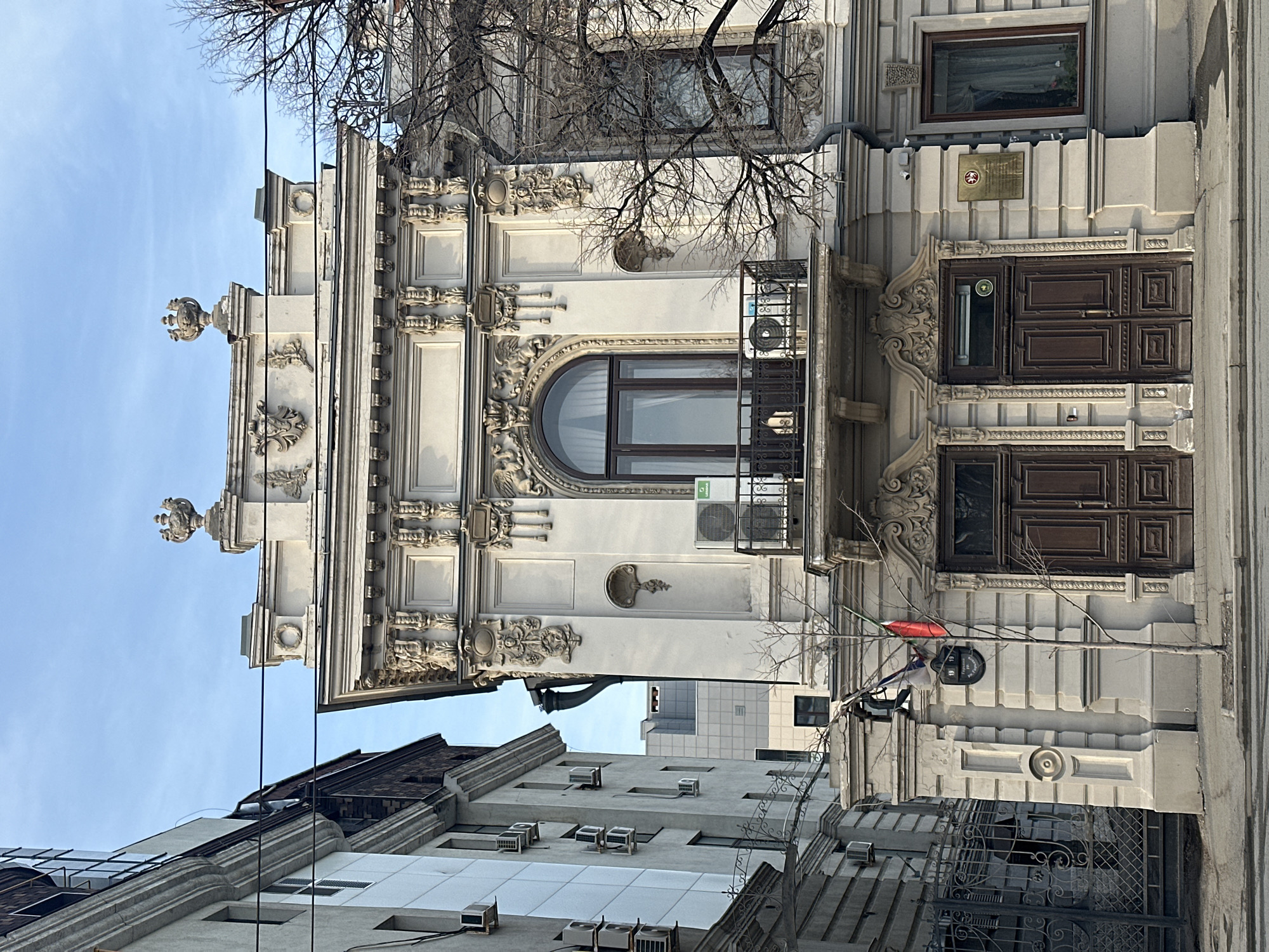 An ornate, three-story building, identified as the House of Ushkov and currently housing the National Library of the Republic of Tatarstan, is depicted on Kremlyovskaya Street, 33, in Kazan, Russia. The building's facade is light-colored, featuring extensive classical relief sculptures, moldings, and decorative elements.
The ground floor presents two large, dark brown wooden double doors with intricate carvings and ornate hardware. Above the right door, a brass plaque with Cyrillic text, indicative of the National Library, is affixed to the wall, with a small security camera mounted above it. To the left of the left door, a small, bare deciduous tree stands on the paved sidewalk. Hung on the wall next to the tree is a dark oval sign with "19 19" and "ГАЛЕРИЯ АРТ" (Art Gallery) in white text, below which two small flags, one Russian (white, blue, red stripes) and one Tatarstan (green, white, red stripes), are draped.
The first floor features a central arched window with dark brown frames, partially obscured by white curtains visible from within, fronted by a wrought-iron balcony. Two air conditioning condenser units, one white and one with a green front panel, are mounted on the facade below this window, flanking the balcony. A decorative shell-shaped niche is carved into the facade to the left of the arched window.
The top floor (second floor) is topped with an elaborate parapet featuring two symmetrical, urn-like finials at the corners and detailed relief carvings, including wreaths and what appear to be mythological figures.
To the immediate left, a taller, plainer, light-colored building with multiple rectangular windows is visible, adorned with numerous external white air conditioning units. A dark, ornate metal gate provides access to a property between the two buildings. To the right of the main building, the bare branches of a deciduous tree partially obscure a portion of an adjacent structure with a dark-framed window. In the background, between the buildings, a modern, grey multi-story building is visible.
The scene is illuminated by daylight under a clear blue sky. No individuals or vehicles are present in the image. The foreground shows a paved sidewalk and a sliver of an asphalt road.