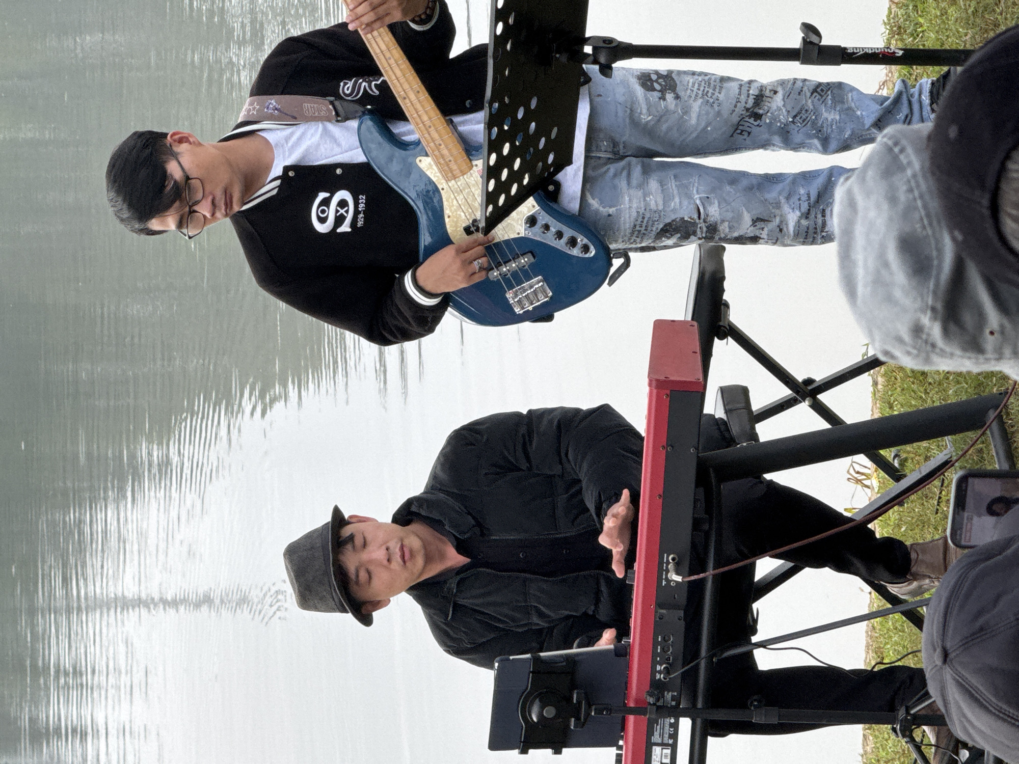 Two men are performing music outdoors by a body of water, possibly Xuan Huong Lake in Da Lat, Vietnam. The man on the left, wearing a black puffer jacket and a grey fedora, is playing a red keyboard, with a tablet mounted on a stand to his left. The man on the right, wearing a black jacket with white trim and light blue ripped jeans with printed graphics, is playing a blue electric bass guitar. Both men are looking down, focused on their instruments. The background features the calm, rippling surface of the water under an overcast sky. In the foreground, out of focus, are audience members, one holding up a smartphone. The scene appears to be a casual outdoor concert or performance.