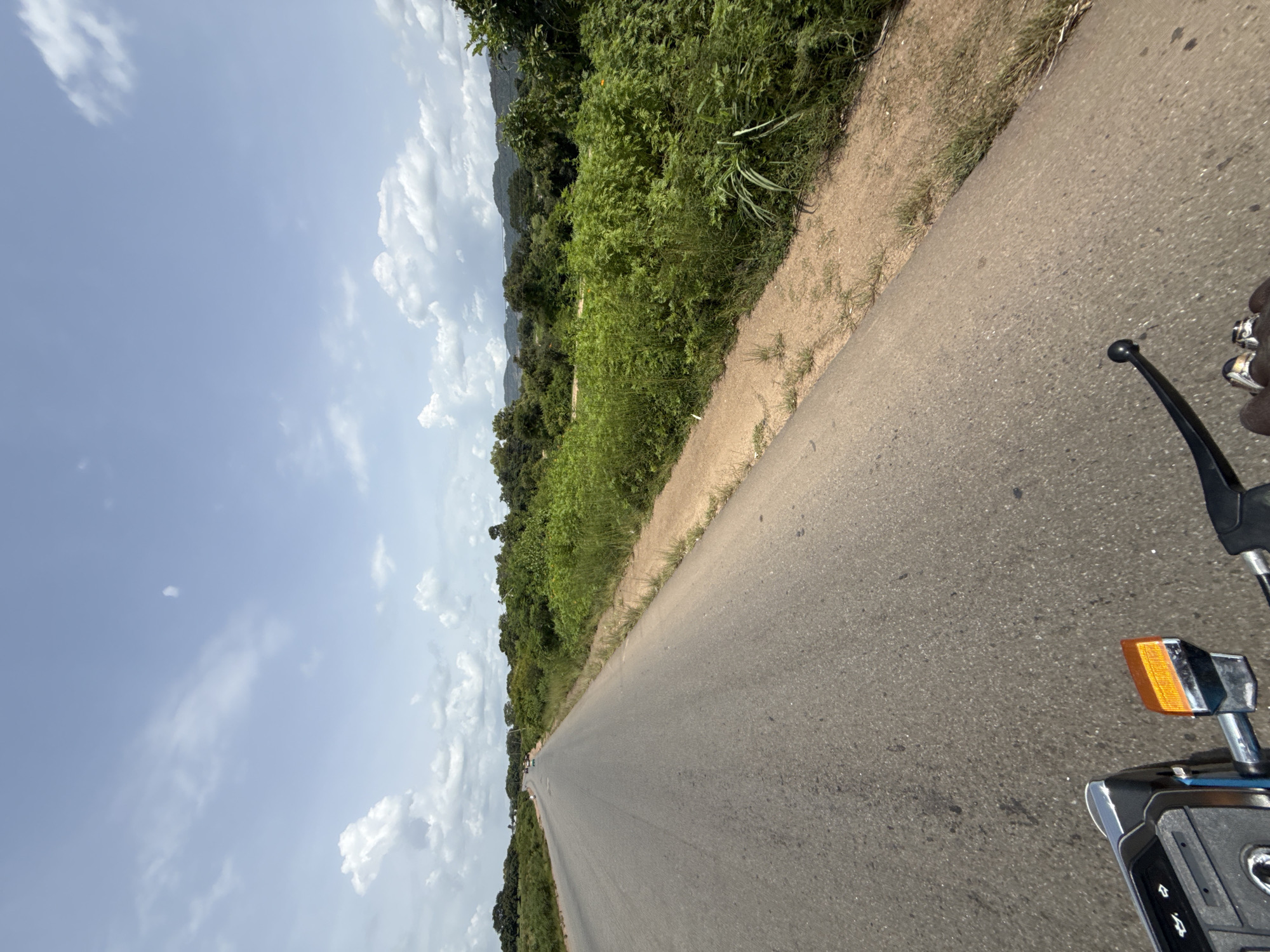 The image shows a view from a motorcycle traveling on a paved road in Tilde, Nigeria. The road stretches into the distance, bordered by lush green vegetation and trees on the right. In the background, rolling hills and a hazy sky with scattered clouds are visible. The foreground of the image includes the handlebars and part of the dashboard of the motorcycle, with a hand wearing rings gripping the handlebar. The scene depicts a rural or semi-rural setting, likely a journey through the Nigerian countryside.