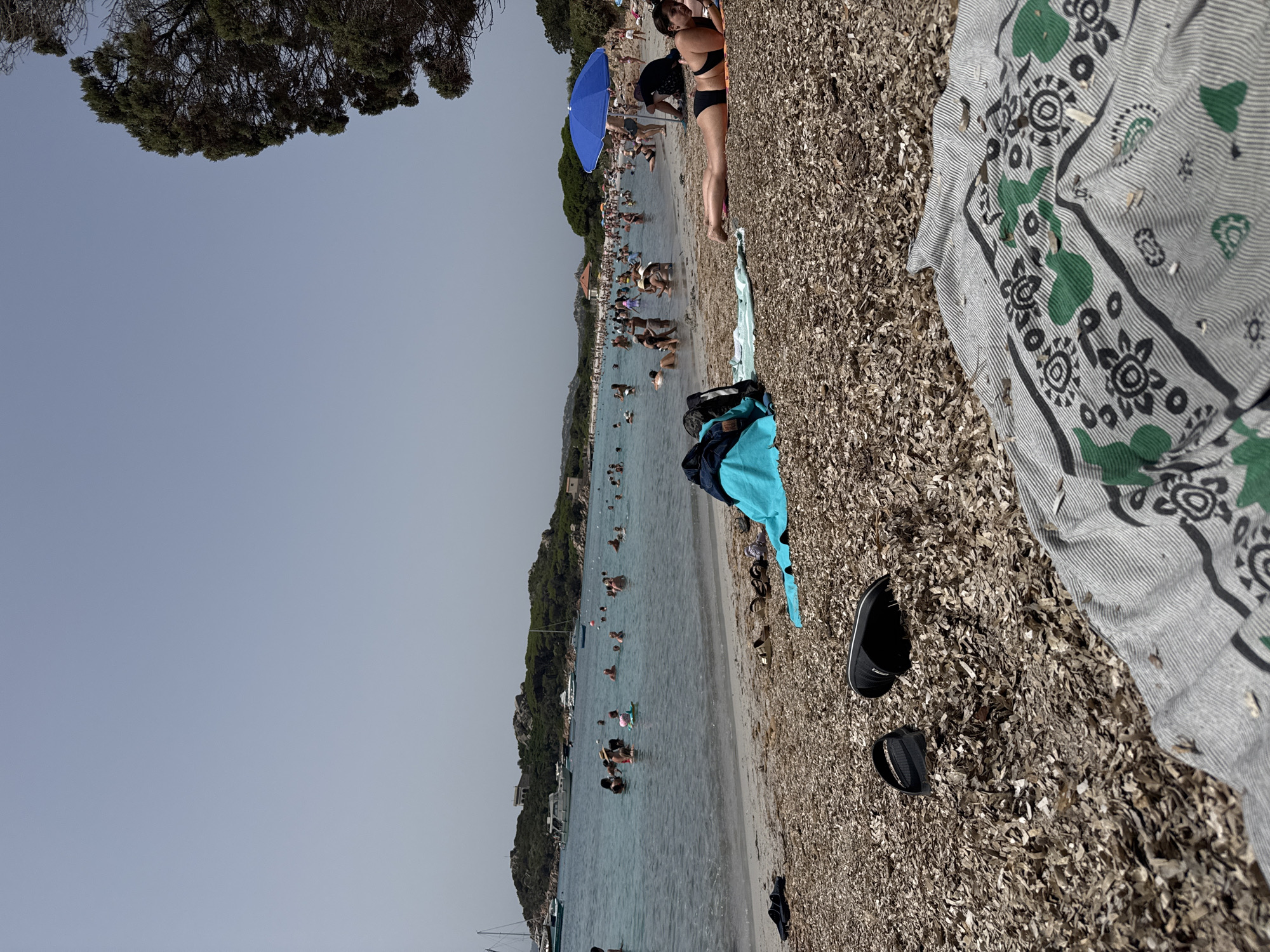 The image captures a sunny day at a crowded beach on the island of Caprera, part of the La Maddalena archipelago in Italy. The foreground is dominated by a beach covered in dried seaweed and pebbles, with several pairs of flip-flops and a folded blanket with a green and grey striped pattern. A bright blue umbrella is visible further up the beach, providing shade to a group of people.
In the middle ground, numerous people are enjoying the clear, turquoise water. Some are swimming, others are standing or wading in the shallow areas. A small pink inflatable toy is in the water. Boats, including sailboats and motorboats, are anchored or sailing in the bay. The background features a hilly, green landscape with sparse buildings and trees, characteristic of the Mediterranean coast. The sky is a pale blue with a hazy quality. The overall scene depicts a lively beach atmosphere during peak summer season.