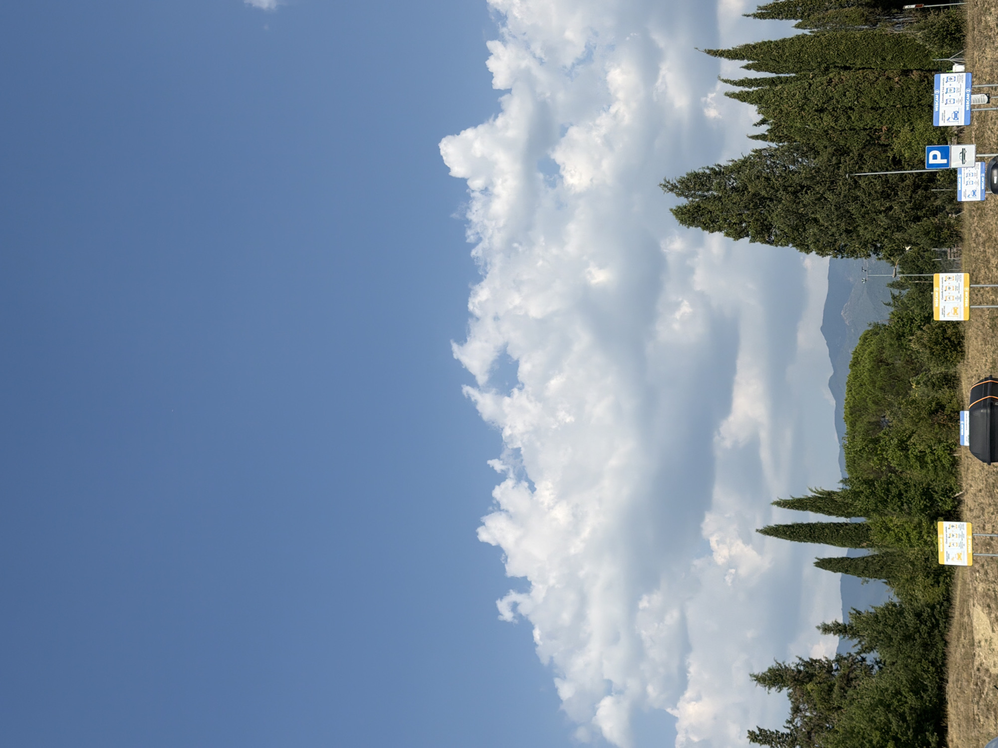 The image depicts a clear blue sky with fluffy white clouds over a landscape of trees and hills. In the foreground, there are several signs. One yellow sign with a diagram and text, and another blue sign with a white parking symbol. A black bin is also visible. The trees are a mix of cypress and other deciduous varieties, and a line of hills is visible in the background. The scene appears to be a roadside or a public area in or near Reggello, Italy, suggested by the style of the signs and the vegetation.