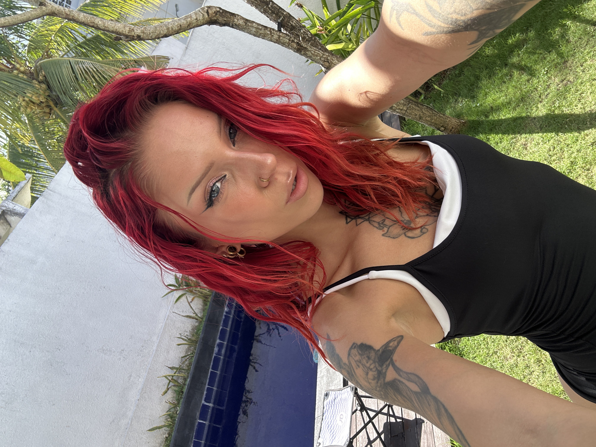 In North Kuta, Indonesia, a woman with bright red hair and blue eyes, adorned with multiple tattoos, poses for a selfie outdoors. She is wearing a black dress with white trim and various piercings, including a nose ring and ear gauges. The background features a swimming pool, lush green grass, and a palm tree, suggesting a tropical or resort setting. Sunlight casts shadows across the scene, indicating it's daytime.