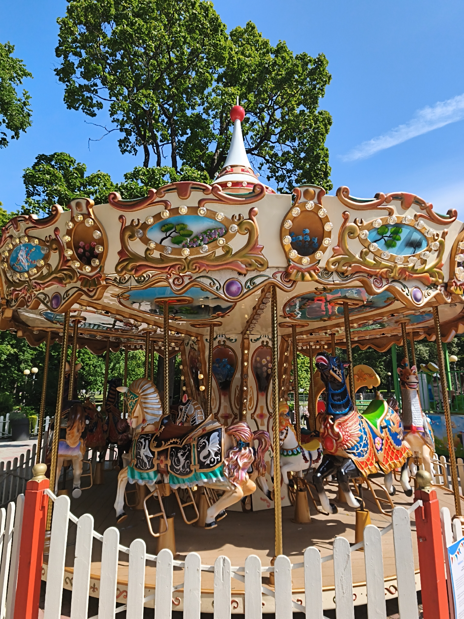 This is a medium-angle, eye-level, outdoor photograph of a carousel in Tallinn, Estonia, on a clear, sunny day. The carousel is the primary subject, occupying most of the frame. It is ornately decorated with cream and gold accents, featuring small, illuminated panels with painted scenes of nature and stylized figures. Several carousel horses are visible, each with unique colors and embellishments; one prominent horse on the right is black with a vibrantly colored, patterned saddle blanket. The carousel is encircled by a white picket fence with red posts. In the background, lush green trees suggest a park-like setting, with a bright blue sky above. There are no visible people interacting with the carousel or its surroundings in this particular shot, though other park attractions may be present off-frame.
