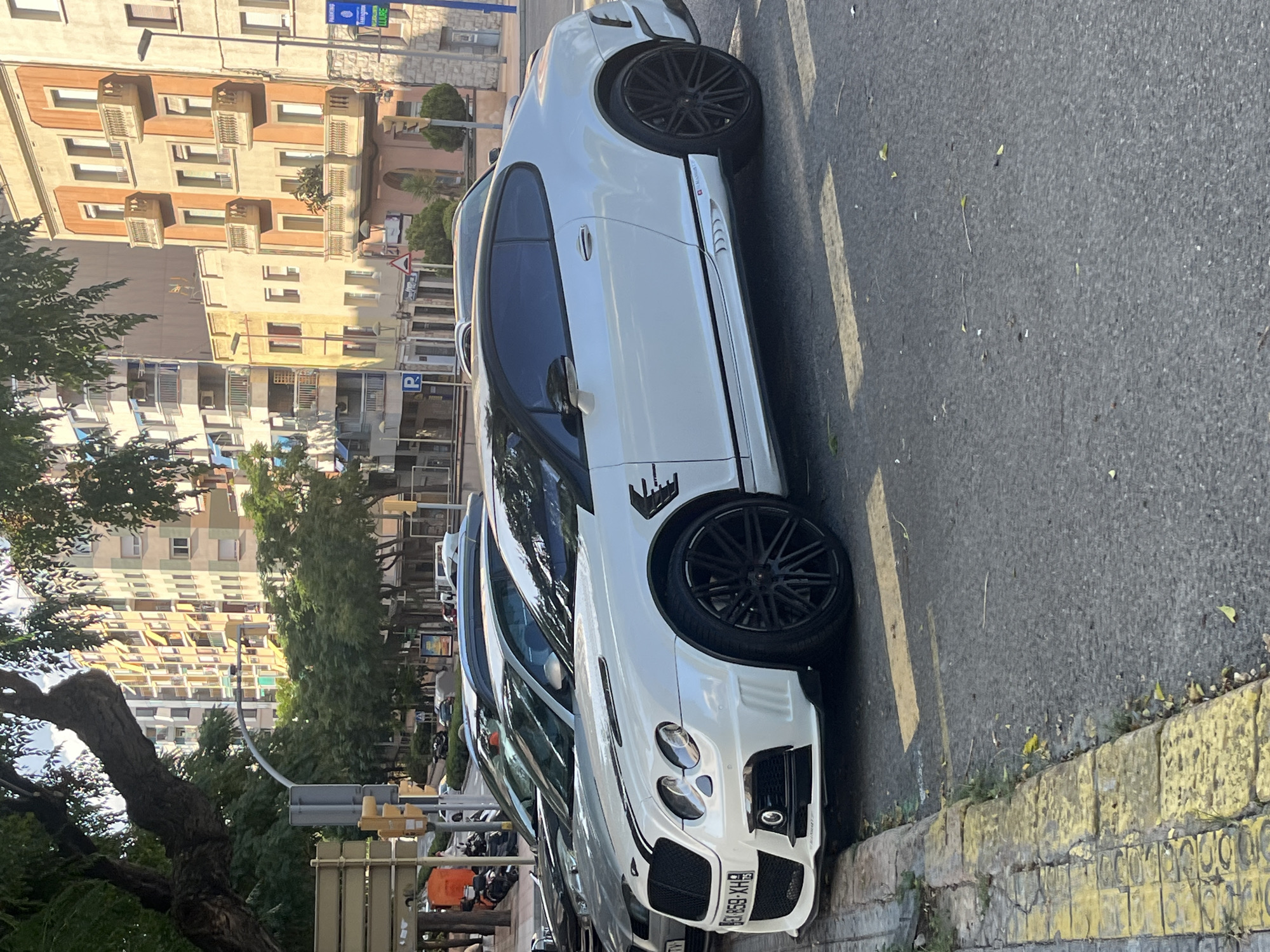 A white Bentley Continental GT Mansory, license plate EX 859 XHB, is parked on a street in Tarragona, Spain. The car has black racing stripes on the hood and a black roof. It is a two-door coupe with tinted windows and large black alloy wheels. In the background, there are apartment buildings with balconies and trees. There are other cars parked behind the Bentley, and a few motorcycles are visible further down the street. The street appears to be made of asphalt, and there is a yellow-painted curb on the left side of the image. A street sign for "Tarragona" and "LLURE" is visible on a pole in the background. The lighting suggests it is daytime, possibly late afternoon, with sunlight casting shadows.